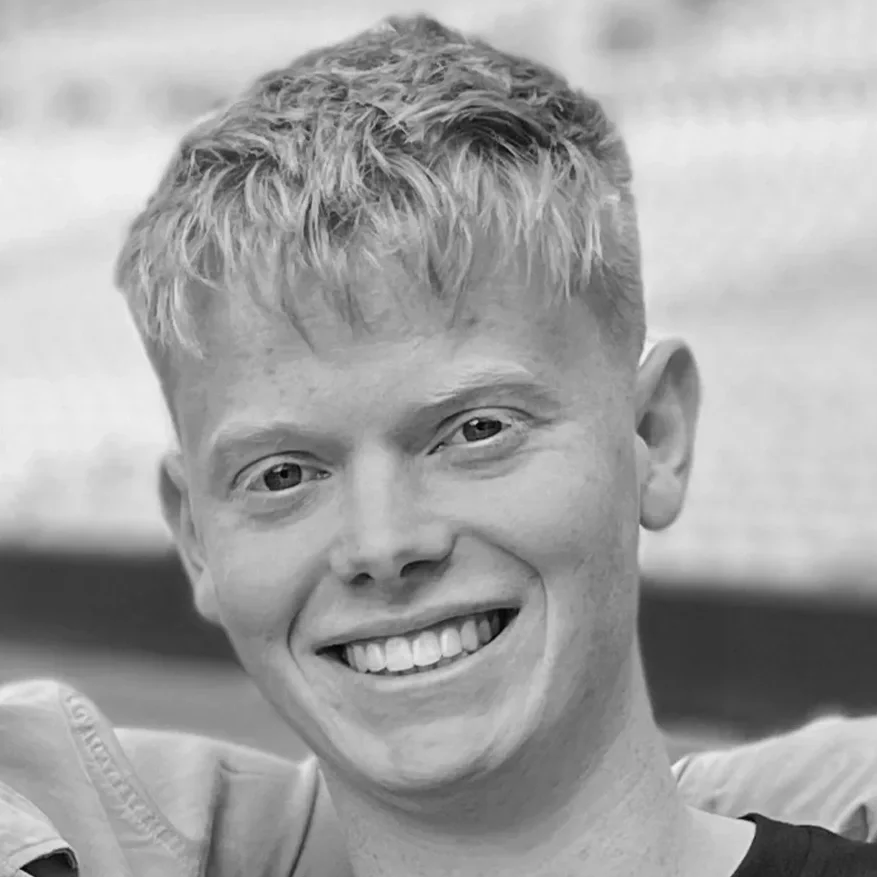 A young man with short, curly hair smiling at the camera, in black and white.