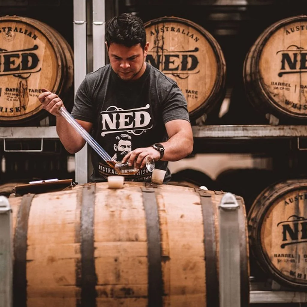 A man in a black T-shirt with the NED logo distills a liquid into a glass using a pipette, surrounded by wooden barrels labeled NED in a warehouse.