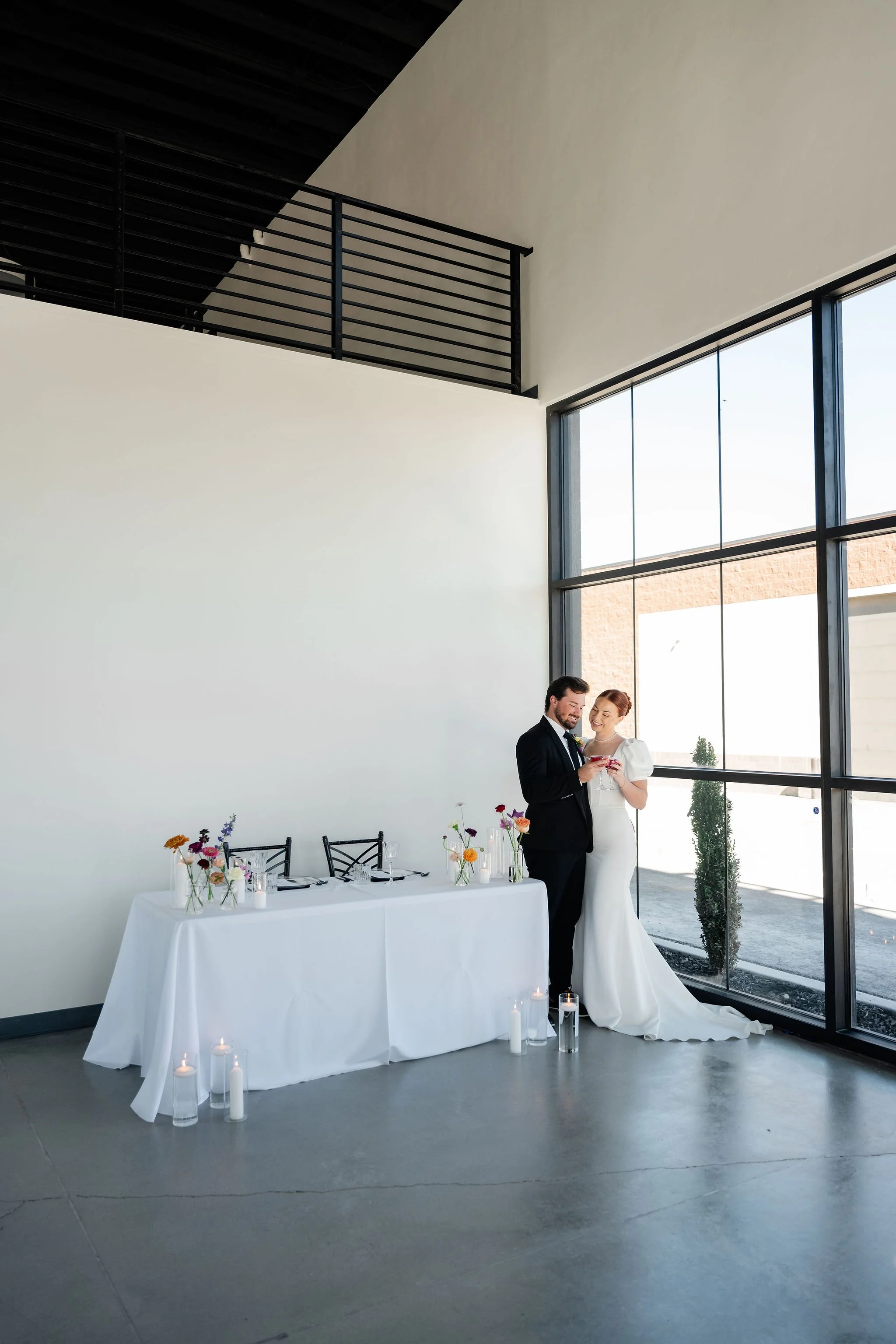 A bride and groom in a black tuxedo and white wedding dress stand close at a bride and groom table in the back section of our event venue's Enchanted Hall
