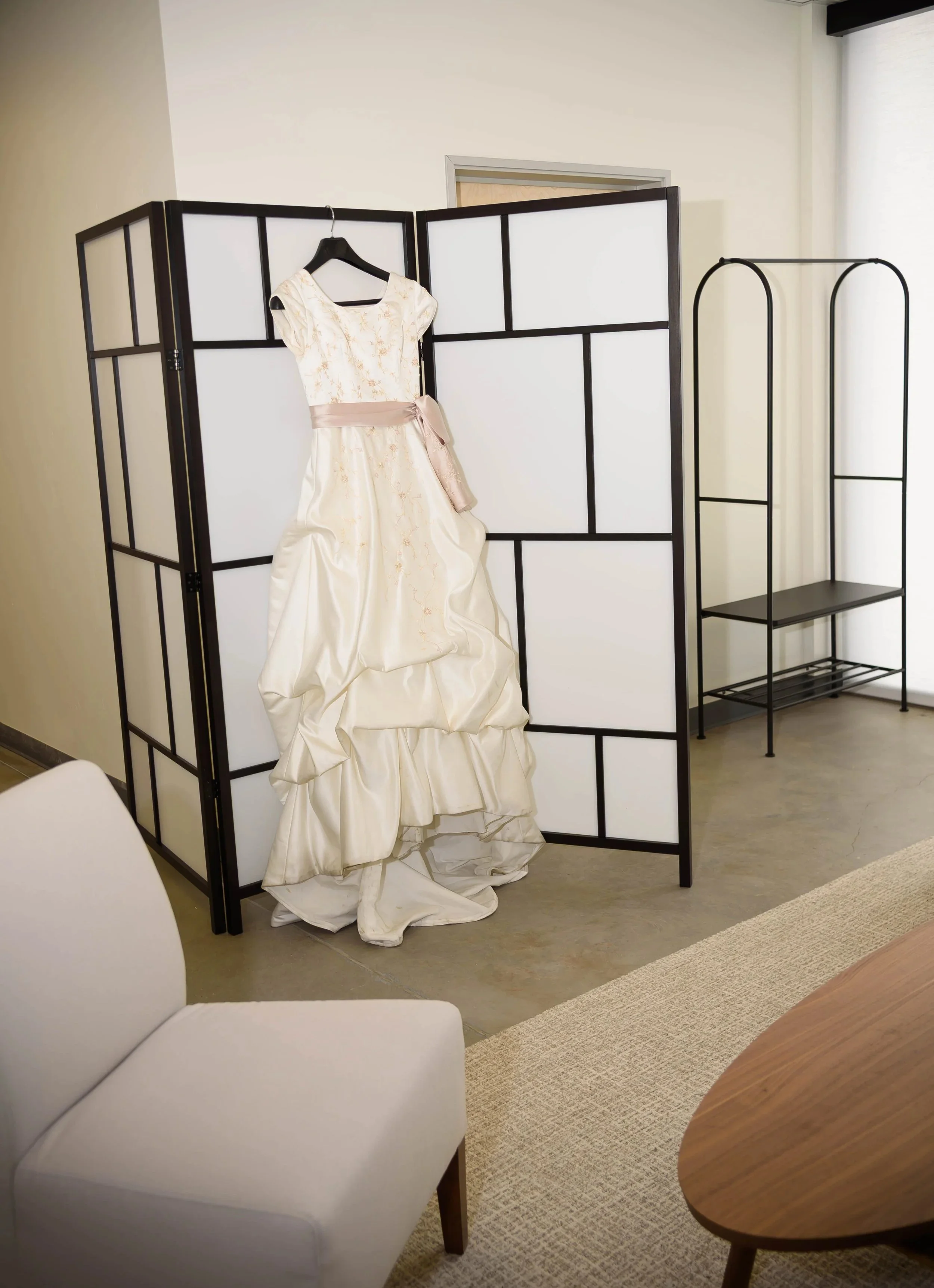 A white wedding gown hung in our getting ready, or prep room, behind a beige chair