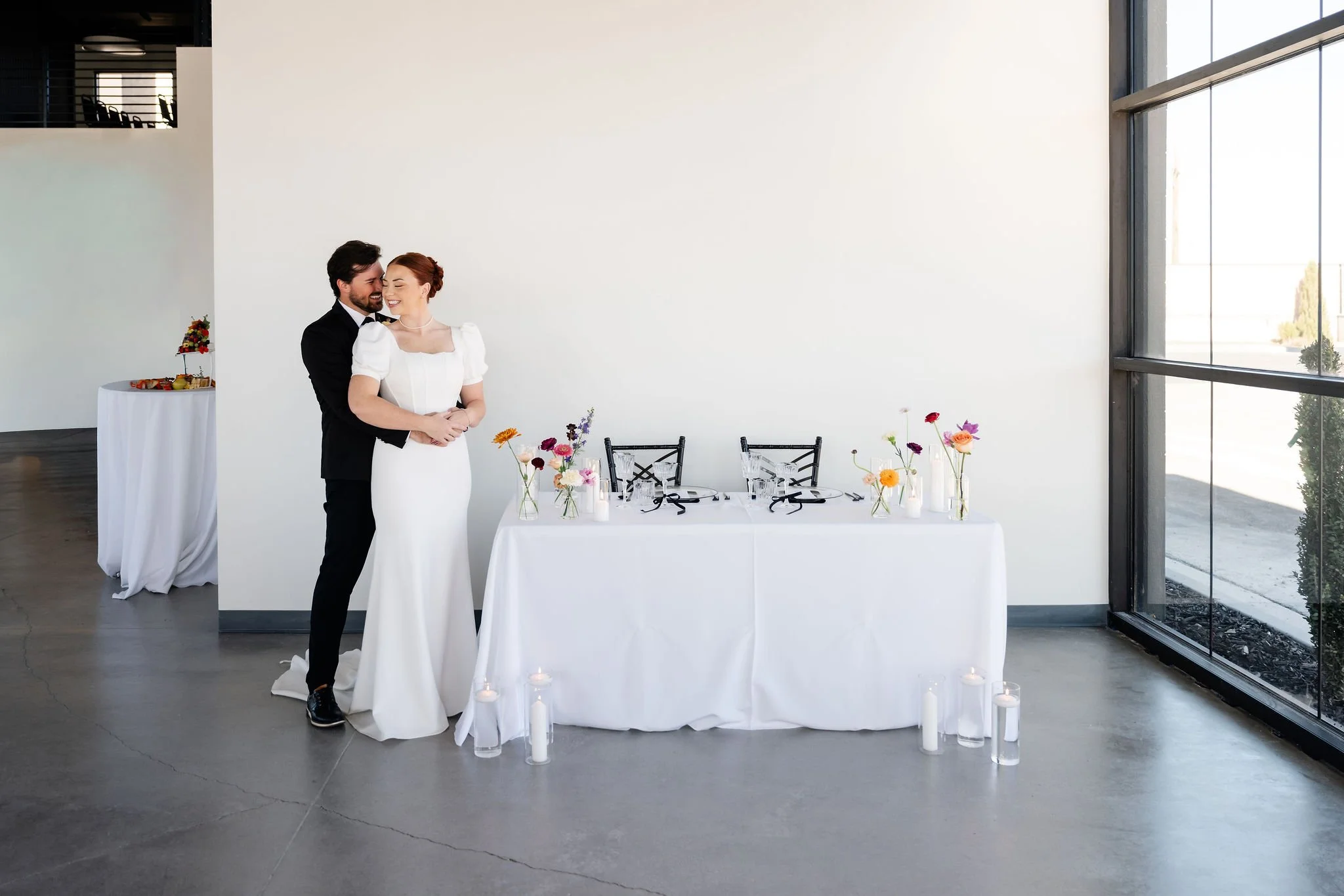 A groom in a dark tuxedo holds his bride in white wedding gown tightly in the back section of our Enchanted Hall near the bride and groom table