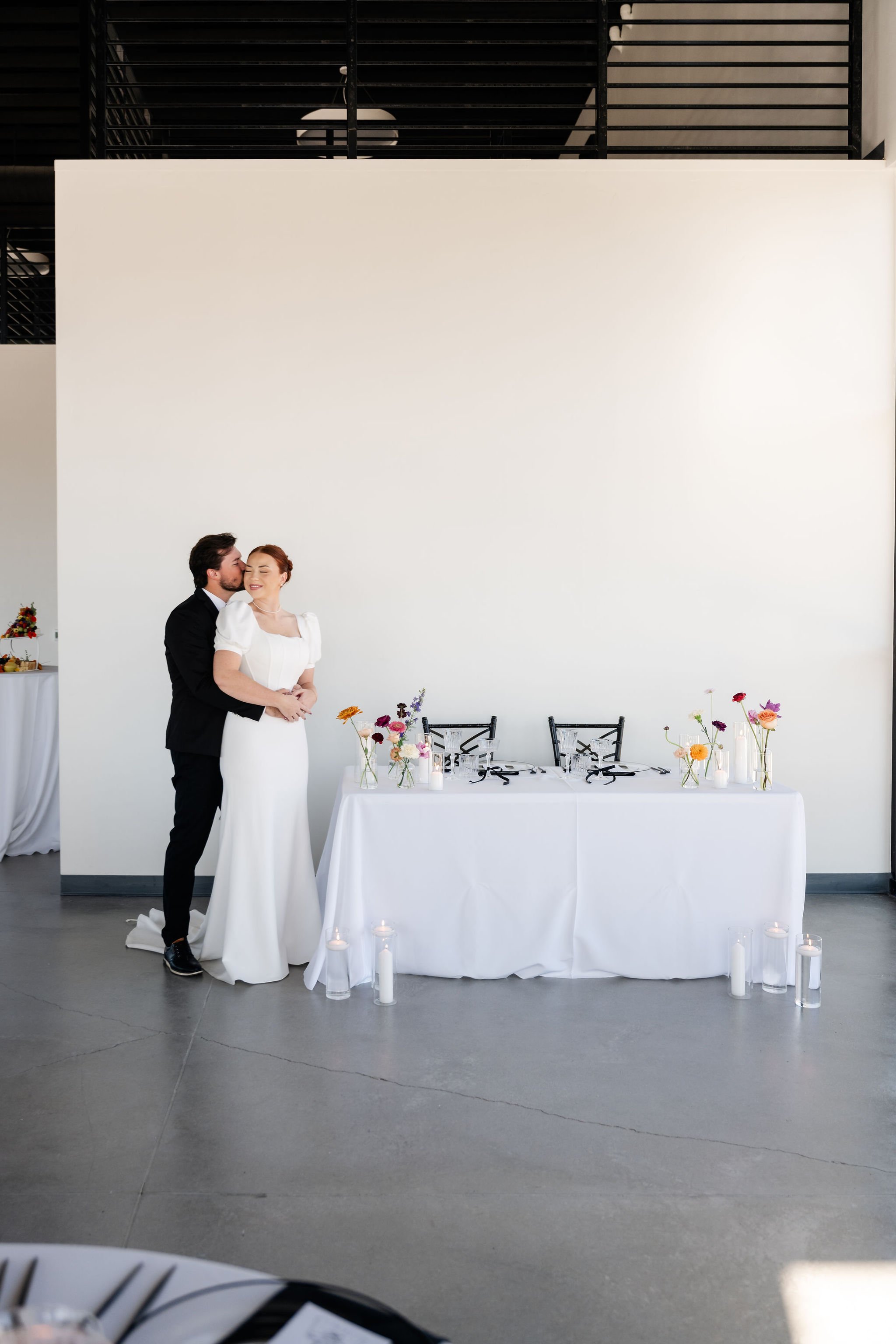 A groom in a dark tuxedo kissed his bride in white wedding gown on the cheek standing behind her in the back section of our Enchanted Hall near the bride and groom table