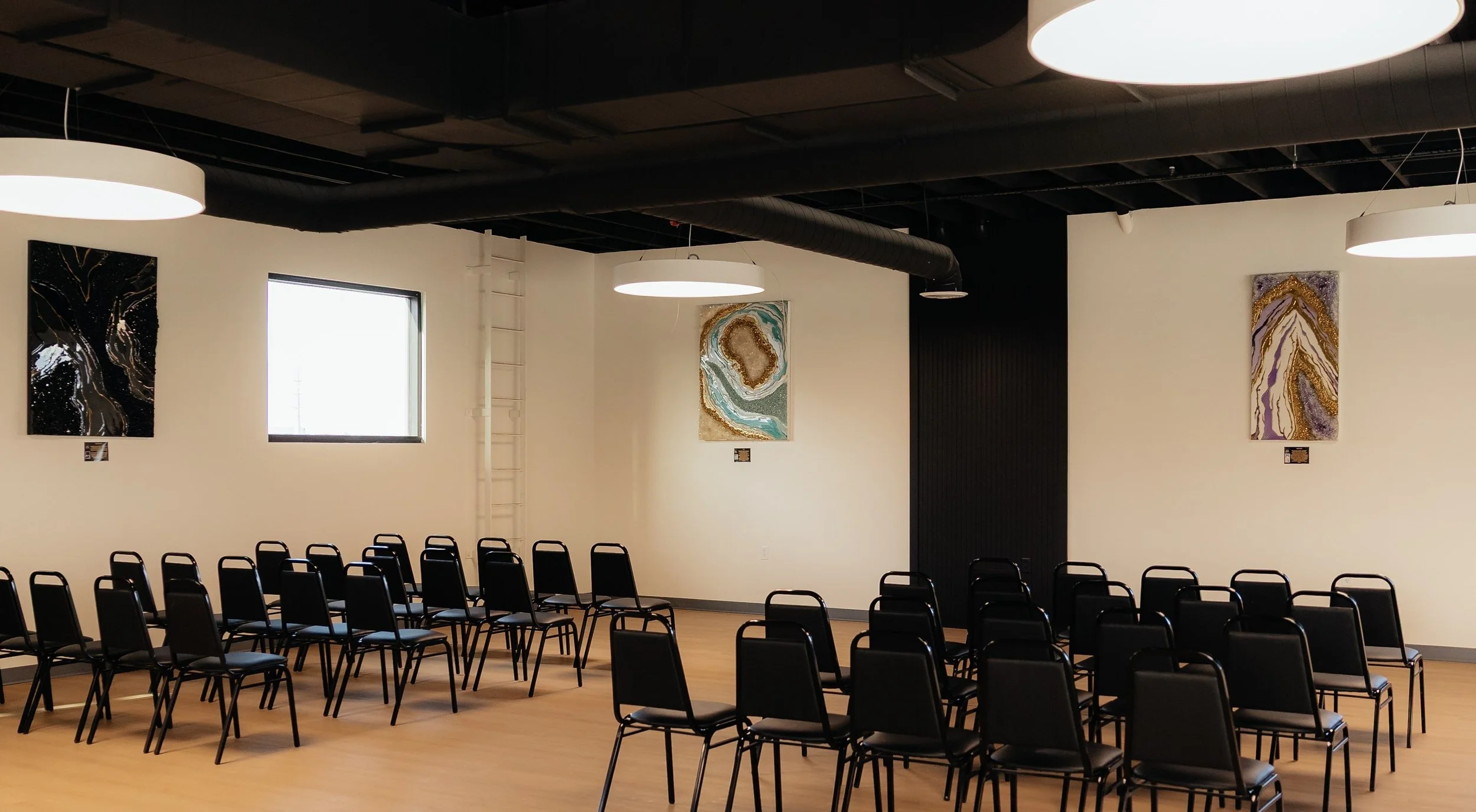 An empty conference or event room with rows of black chairs facing a beige wall. The wall has three abstract paintings and two windows. The ceiling is black with large round white lights and exposed ductwork.