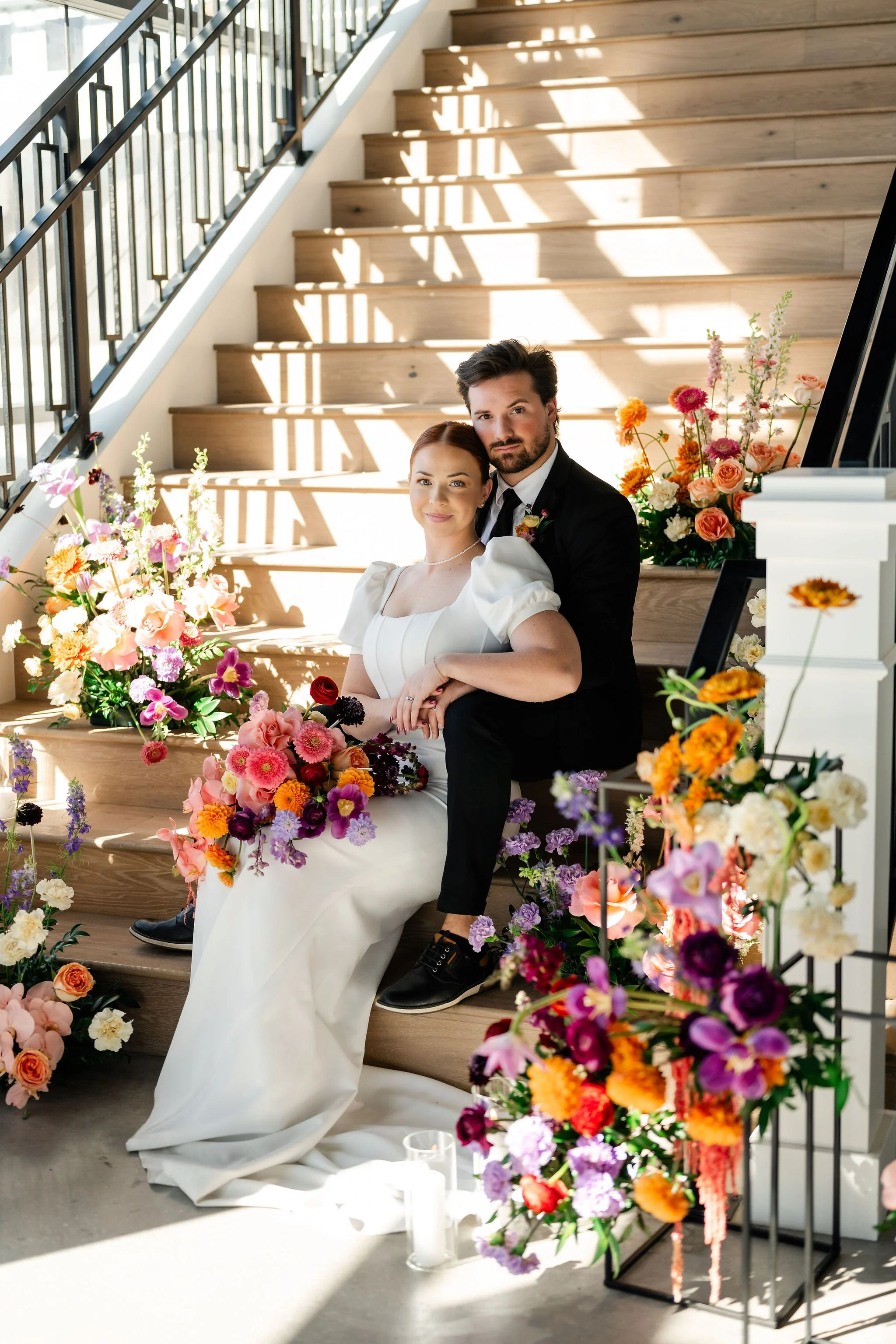 Bridge and groom in white dress and black tuxedo sitting on our grand staircase surrounded by bright colorful flowers