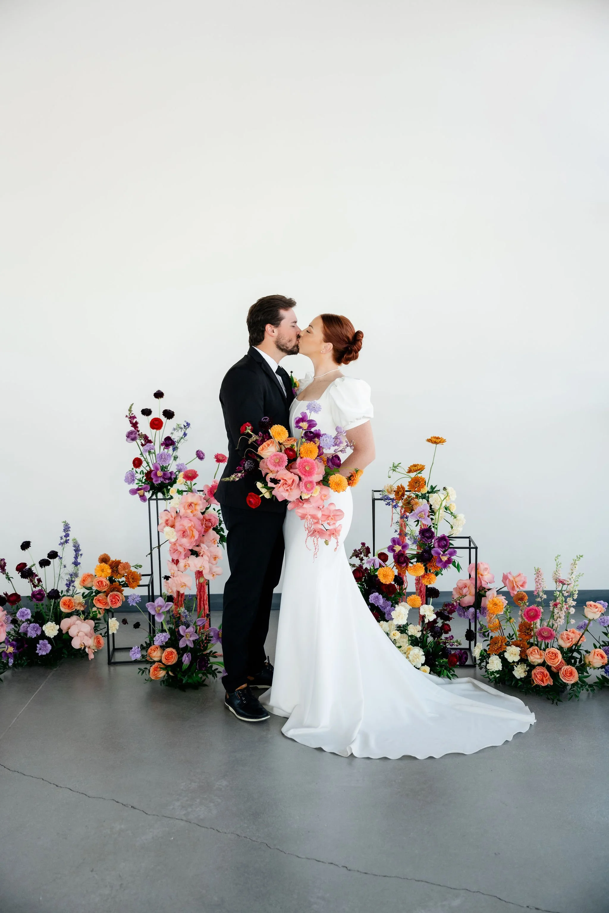 A bride and groom in a black tux and white wedding dress kiss in front colorful floral arrangements set up as the centerpiece of a ceremony aisle