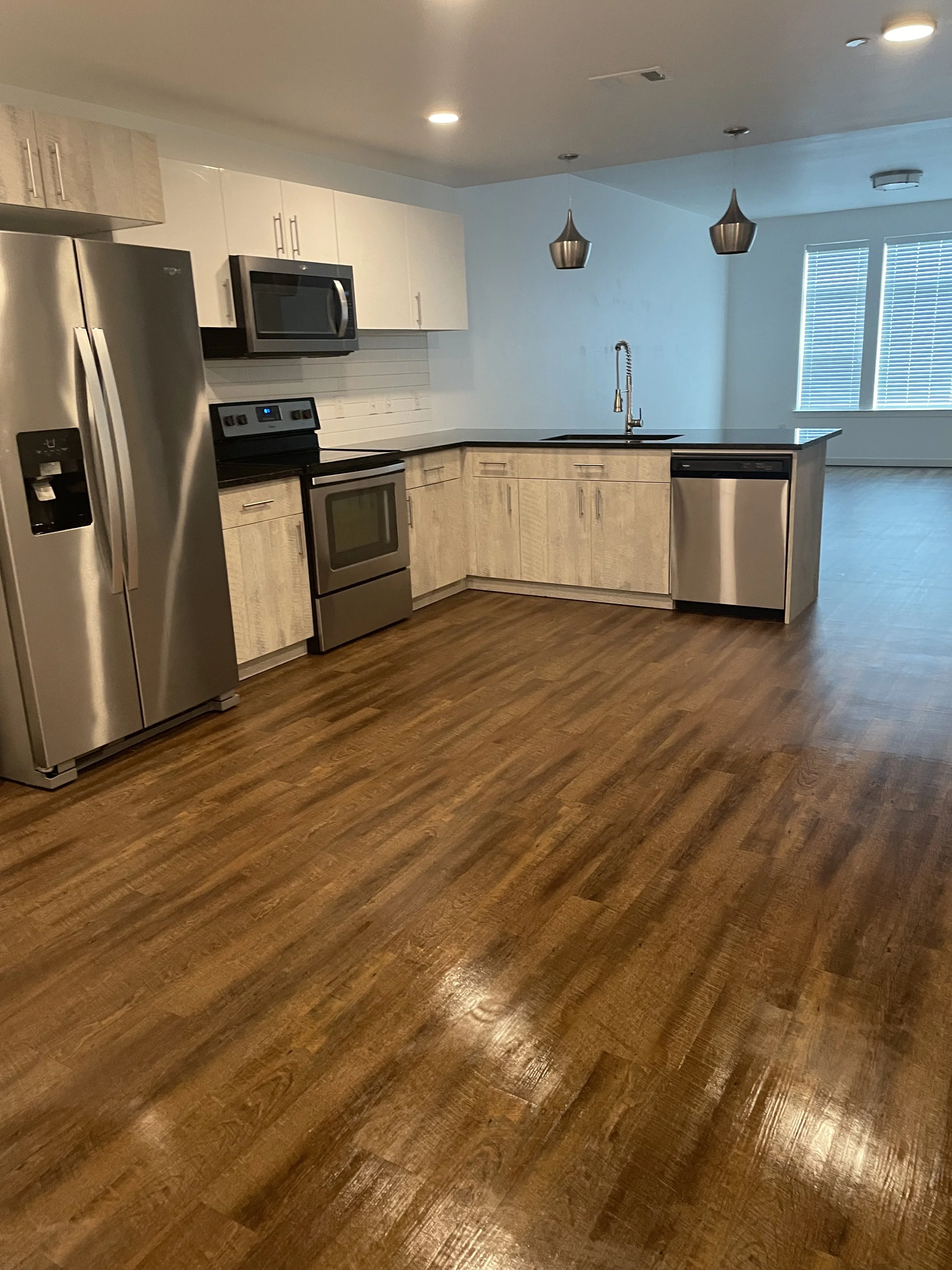 Modern empty kitchen with stainless steel appliances, wood flooring, white upper cabinets, light wood lower cabinets, pendant lights, a window with blinds, and an open layout.