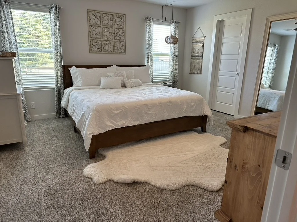 A cozy bedroom with a large bed made with white bedding, flanked by two windows with patterned curtains. There is a wooden dresser to the right, a mirror reflecting part of the room, and a plush cream-colored rug on the carpeted floor. Decorative art hangs on the wall.