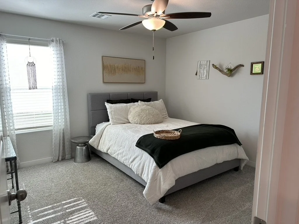 A bedroom with a gray bed, white bedding, a black throw blanket, and plush pillows. There is a window with sheer curtains and decor on the white wall, including a framed picture, a wall sculpture, and a small square green art piece. A ceiling fan with a light is above.