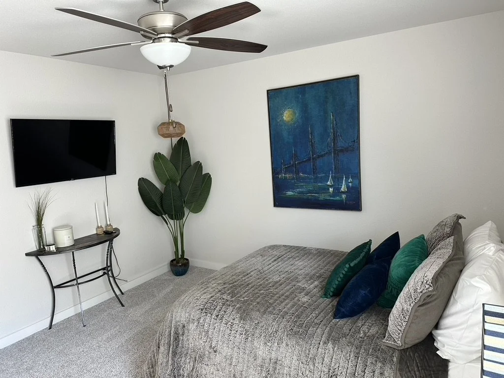 A bedroom with a bed covered in a gray textured blanket and multiple pillows in various shades of green and blue. A modern ceiling fan with dark wood blades is on the white ceiling. A flat-screen TV is mounted on the white wall next to a large green plant in a pot. On the wall above the bed is a blue painting of sailboats on water under a moonlit sky.
