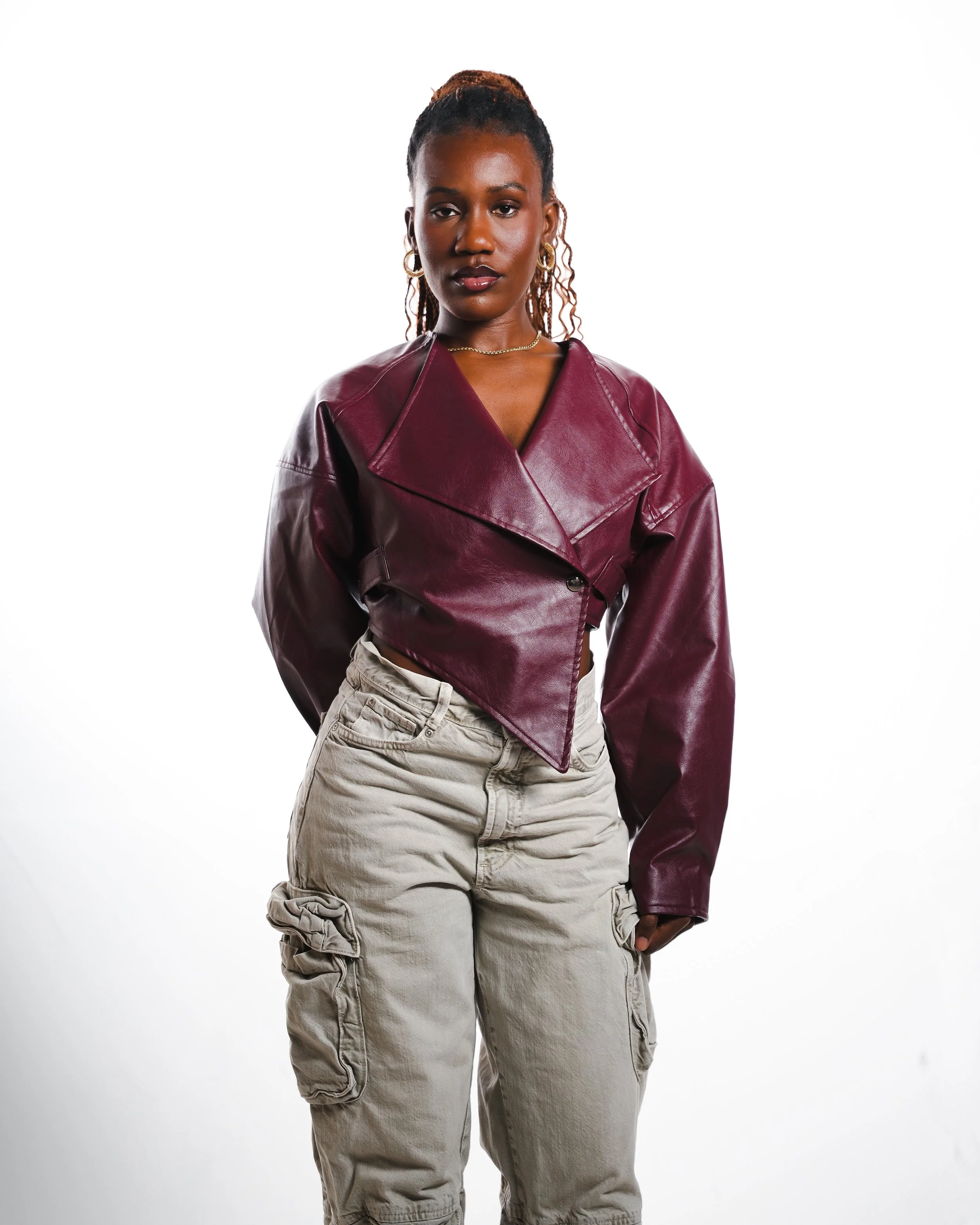 A woman with dark skin and braided hair stands against a white background, wearing a burgundy leather jacket and beige cargo pants.