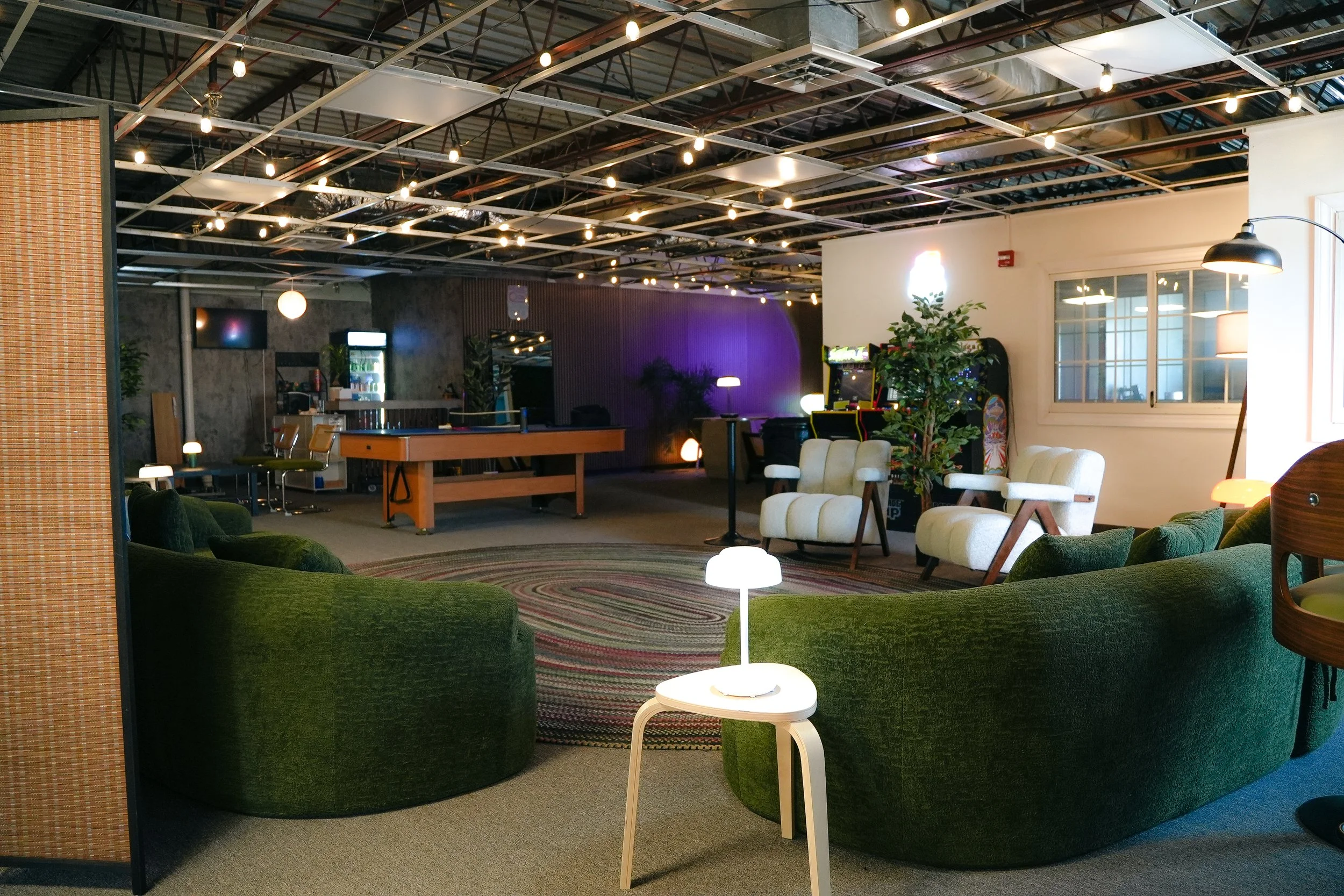 Interior of a stylish lounge or game room with green velvet sectional sofa, white chairs, arcade machines, and a pool table, illuminated by string lights.