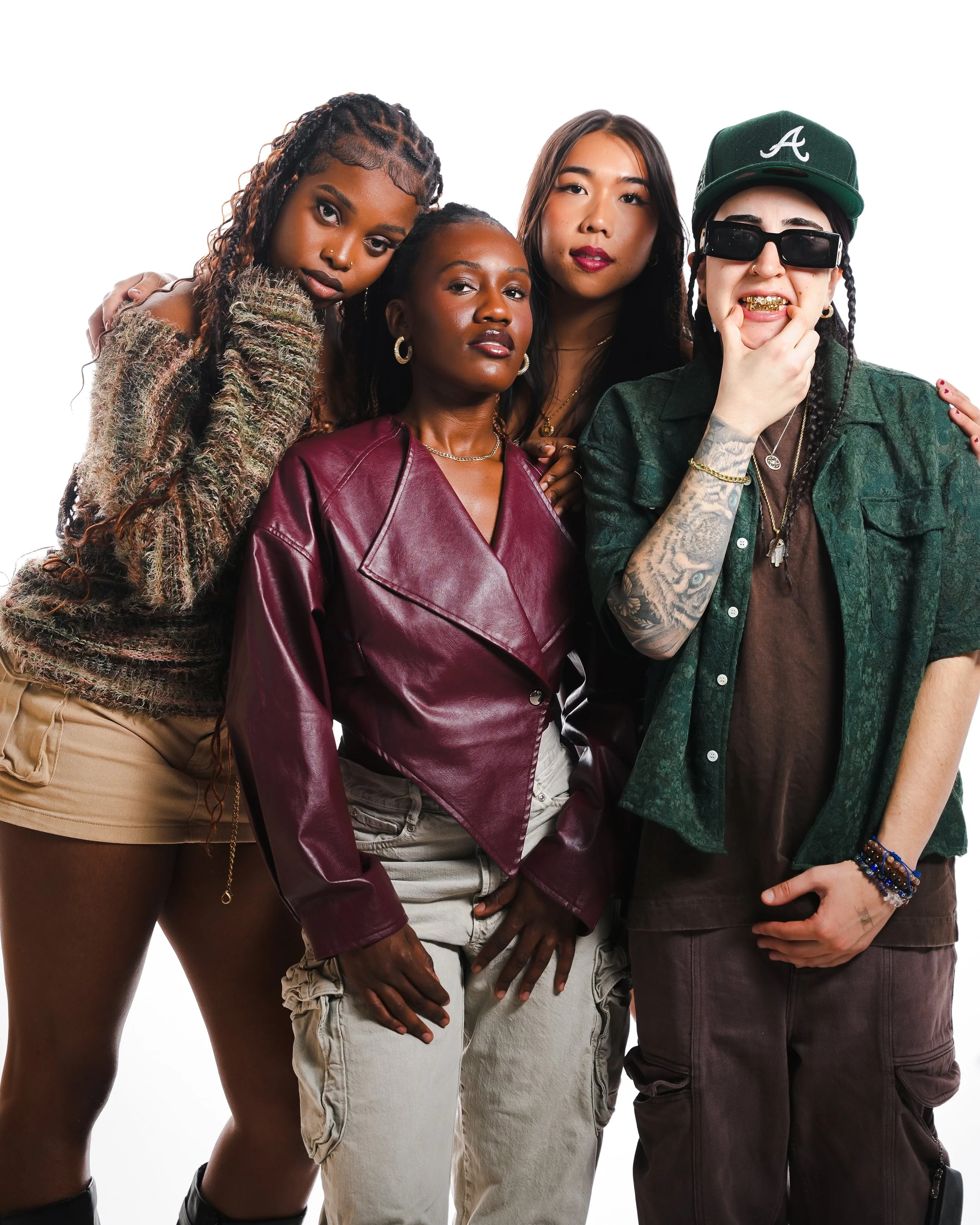 A diverse group of four women posing together, with one woman wearing sunglasses, a green cap, and gold teeth.