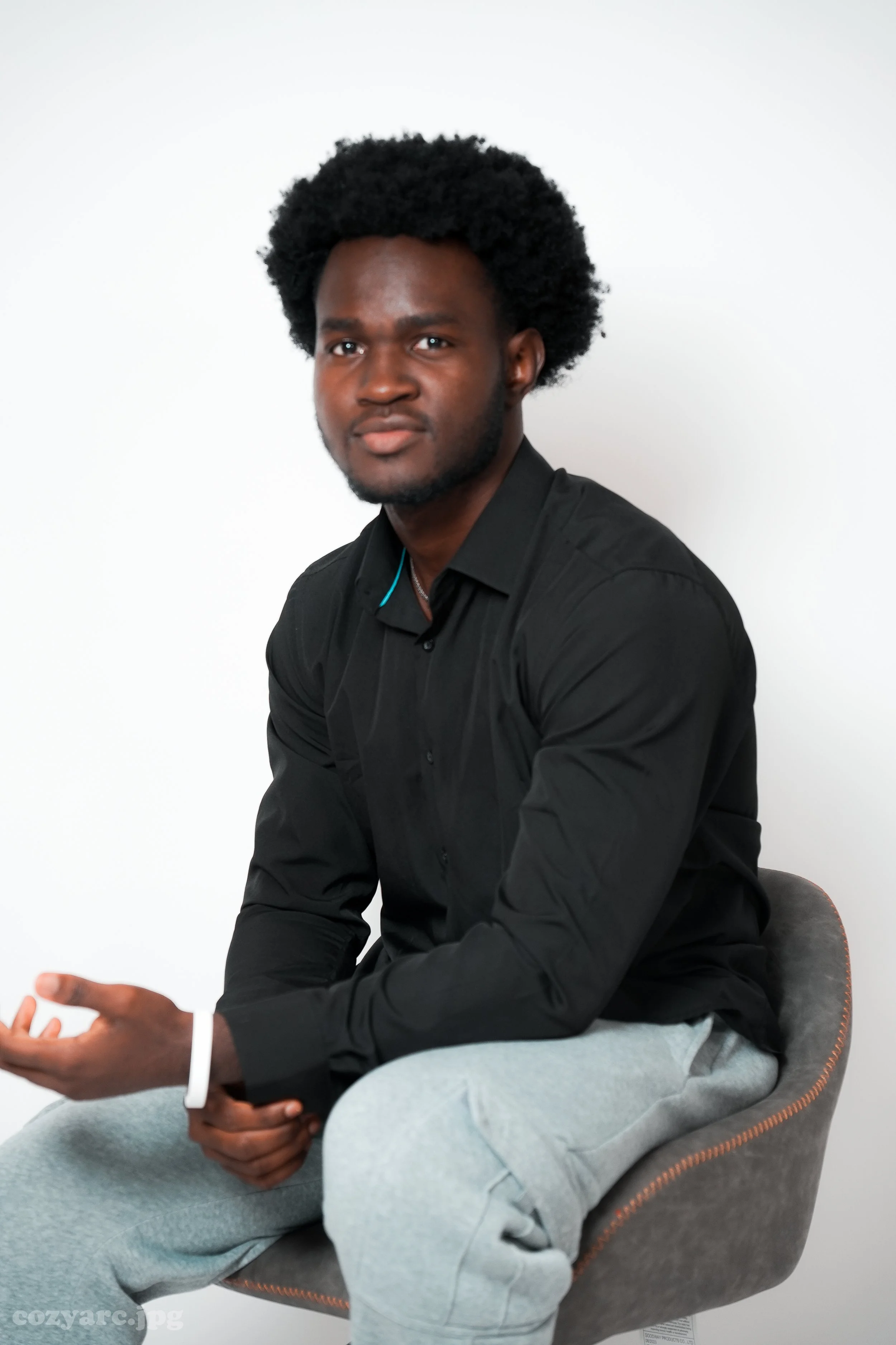 A young man with curly black hair, wearing a black shirt and light gray sweatpants, sitting on a modern gray chair against a plain white background.