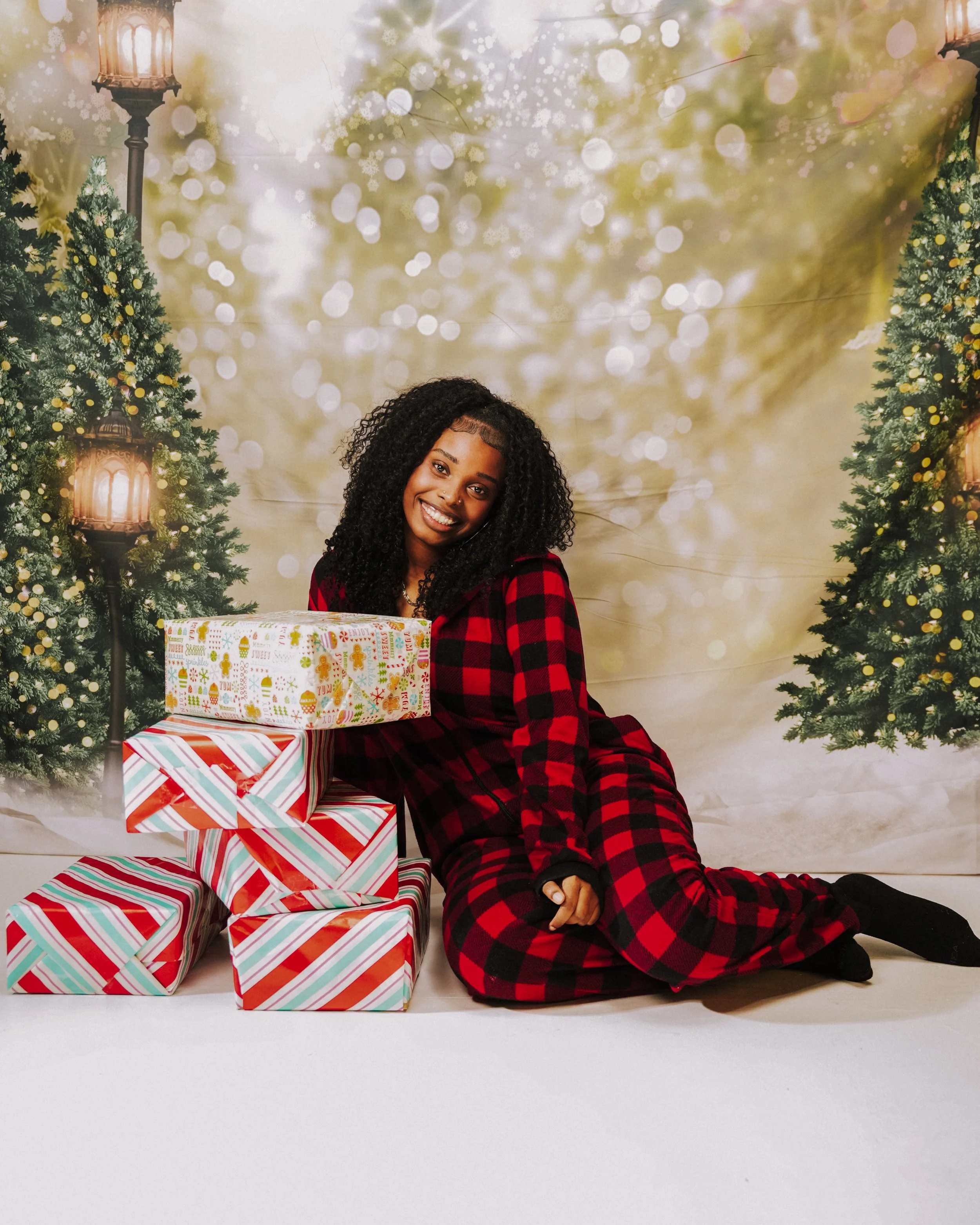 Smiling woman in red and black plaid pajamas sitting on the floor next to a pile of wrapped Christmas presents, with Christmas trees and festive lighting in the background.