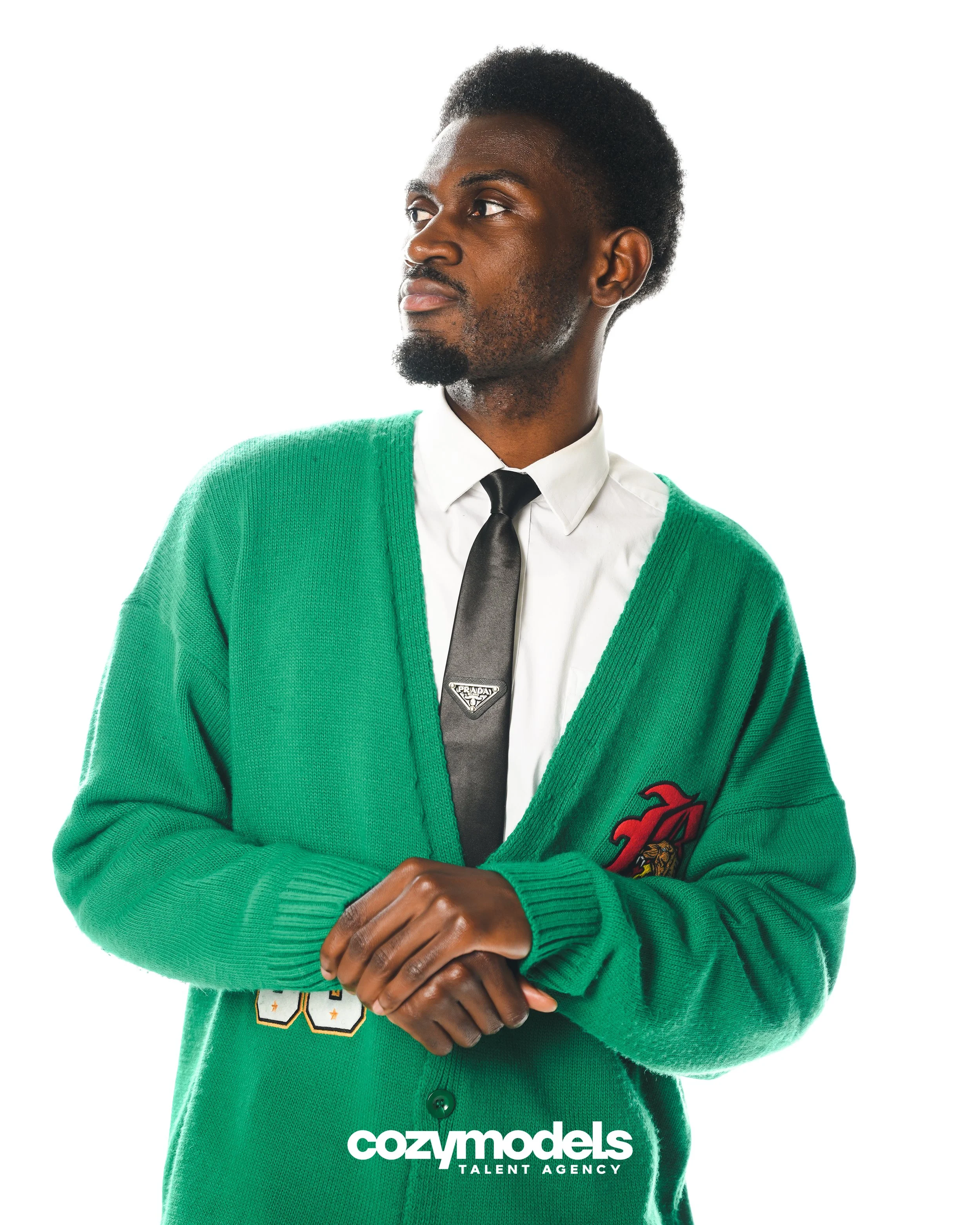 A man wearing a green cardigan sweater with patches over a white shirt and black tie, looking to the side against a white background.