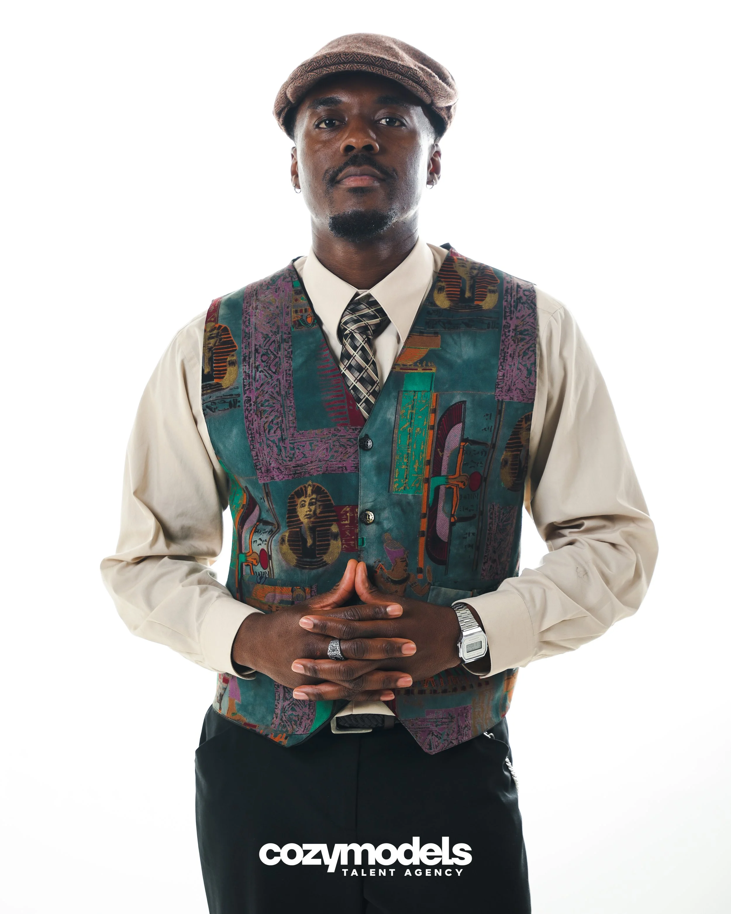 A man wearing a beige shirt, colorful vest, patterned tie, and brown cap poses with hands clasped in front, against a white background with cozymodels Talent Agency logo.