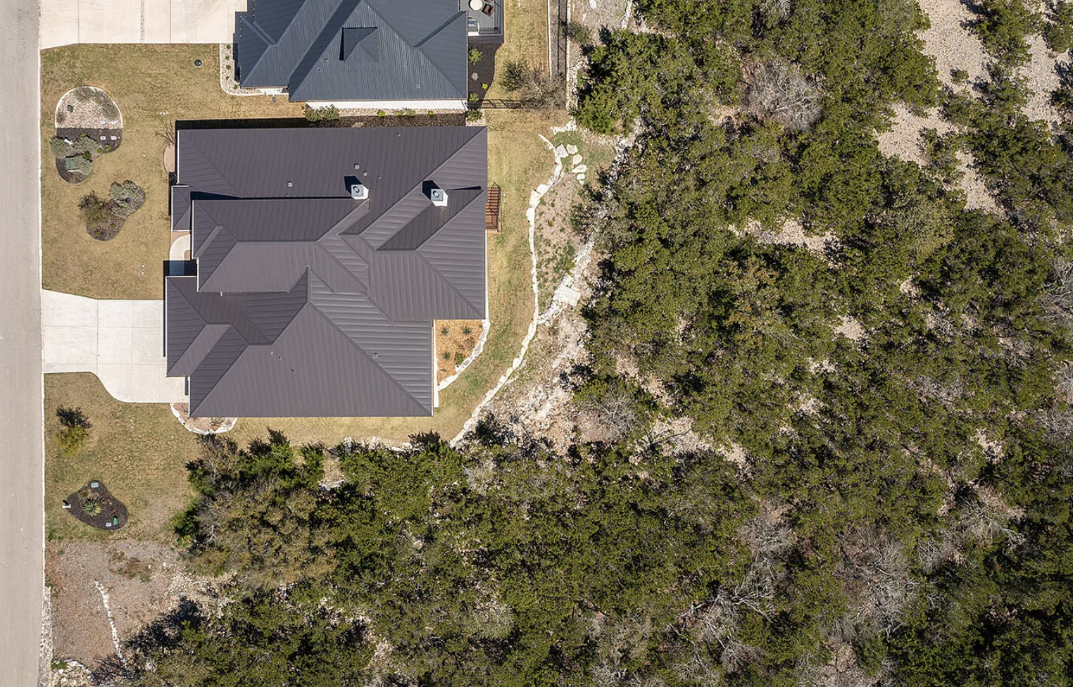 An aerial view of a house with a dark gray metal roof, a driveway, and a landscaped lawn, bordered by a dense forest of trees.