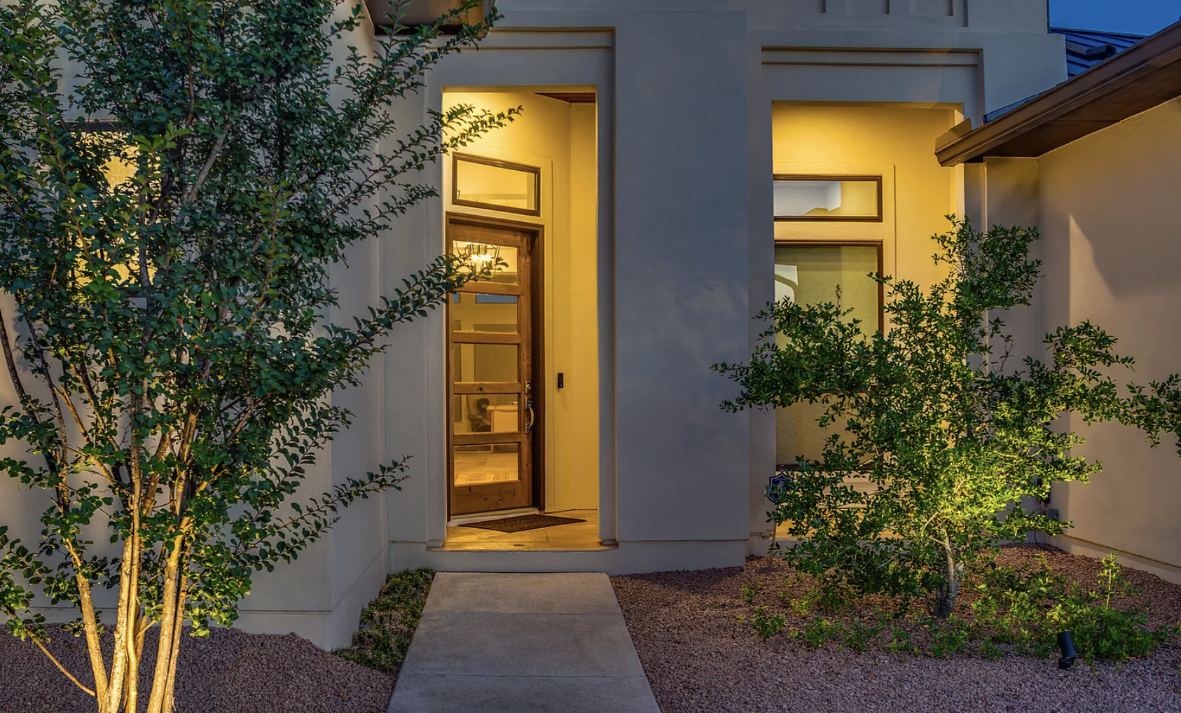 Modern house entrance with lighted doorway, wooden door with glass panel, surrounding windows, green bushes, and a walkway leading up to the door, during dusk or early evening.