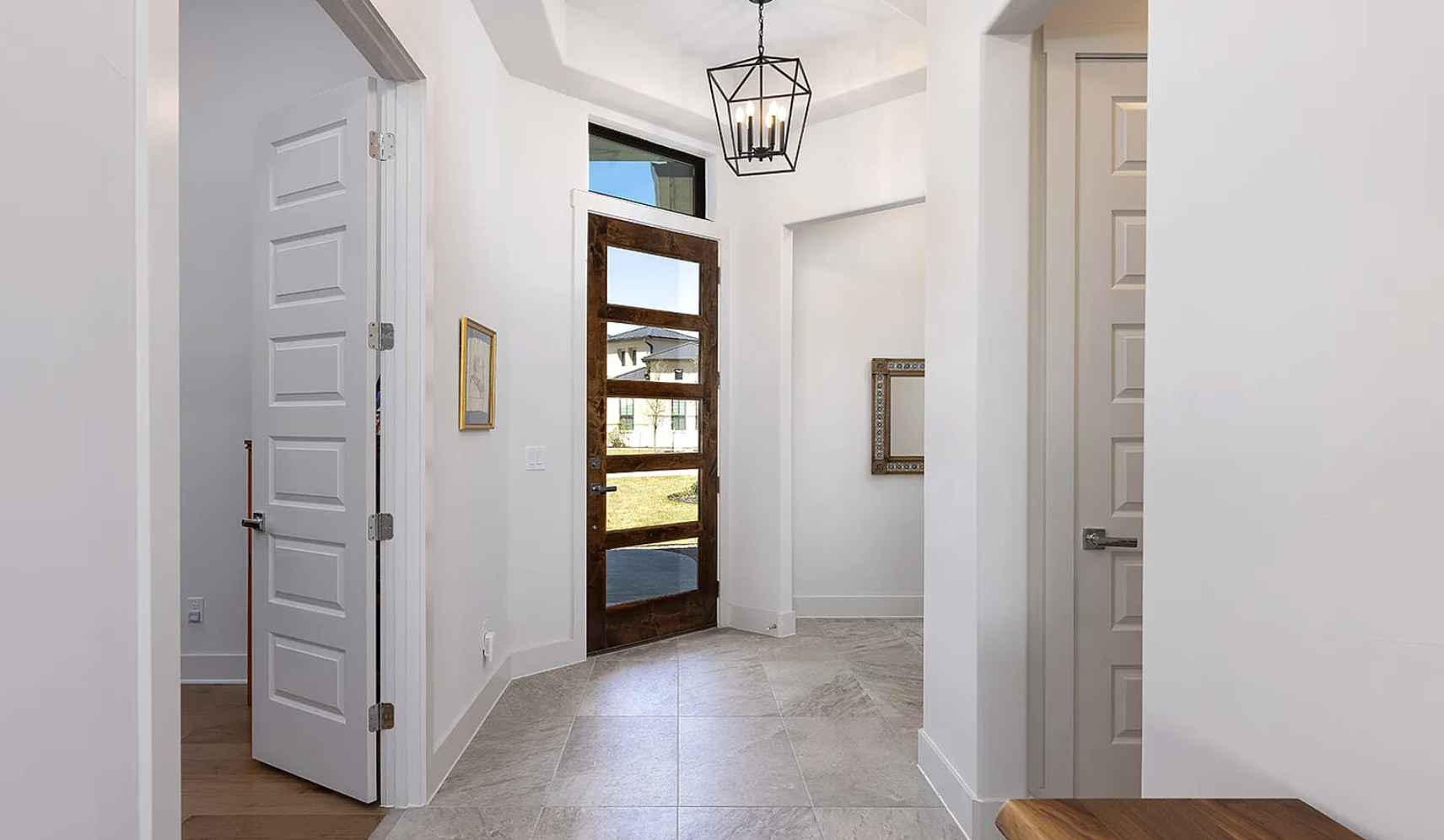 Interior view of a modern home entrance with a wooden front door featuring horizontal glass panels, white walls, tiled flooring, and a contemporary black geometric chandelier hanging from the ceiling.