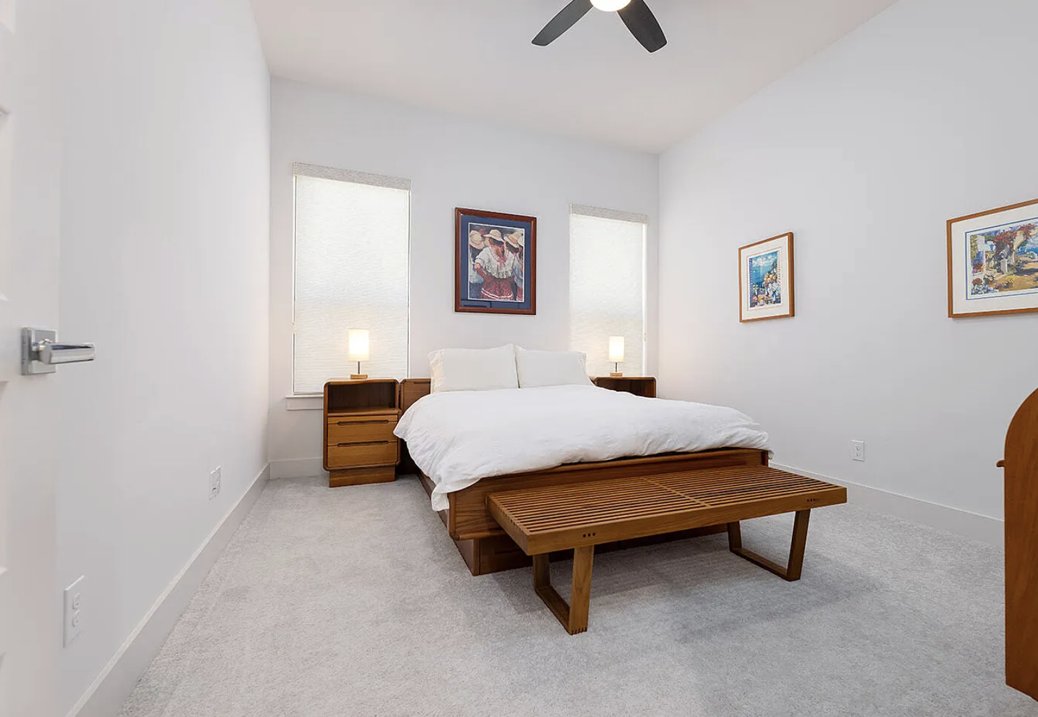 A bedroom with a white bed, wooden nightstands with lamps, a wooden bench at the foot of the bed, and framed artwork on the white walls. Two windows with blinds flank the bed, and a ceiling fan is above.