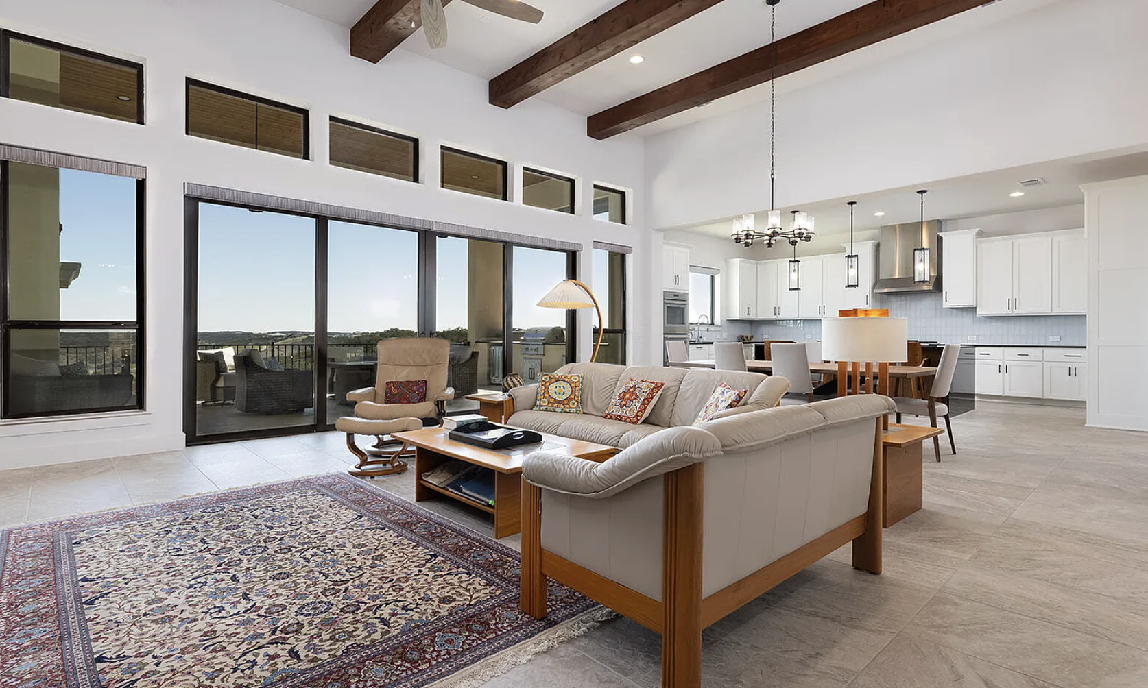 Bright living room with large windows, white walls, wooden ceiling beams, beige sofa with colorful pillows, wooden coffee table, beige armchair, outdoor patio with seating, open white kitchen with dining area, chandelier, pendant lights, and tiled fl