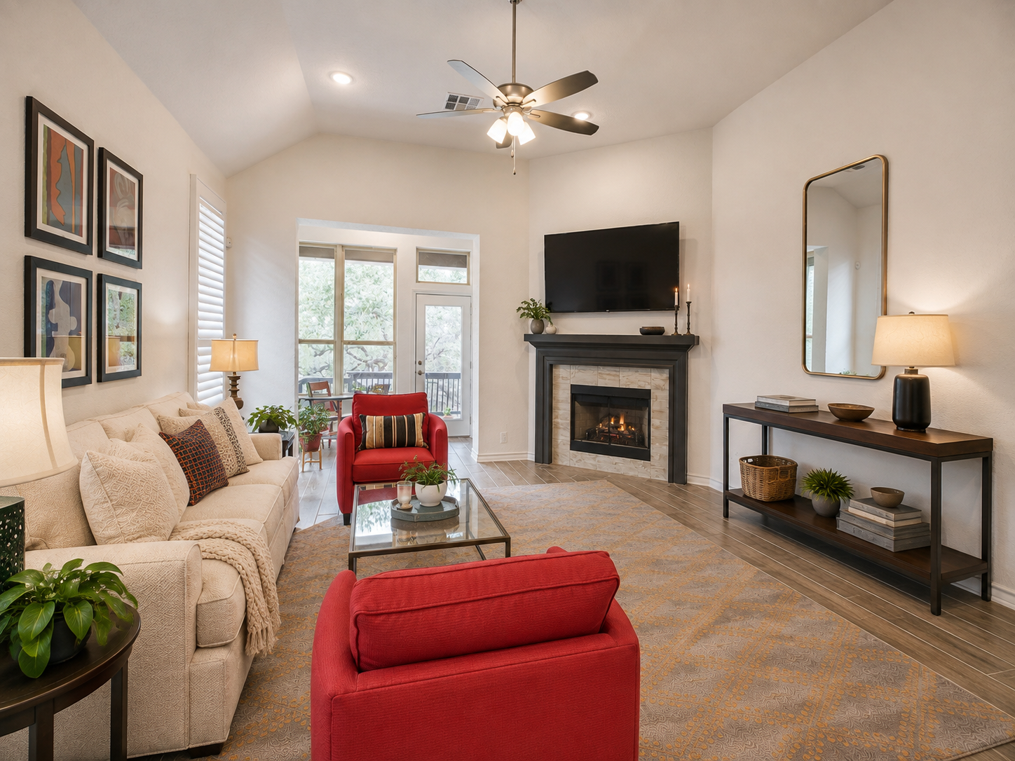 A cozy living room with a beige sofa, red armchairs, a glass coffee table, a fireplace with a mounted TV above it, and a console table with lamps and decorative items of 3051 Blenheim Park, Bulverde, Texas.