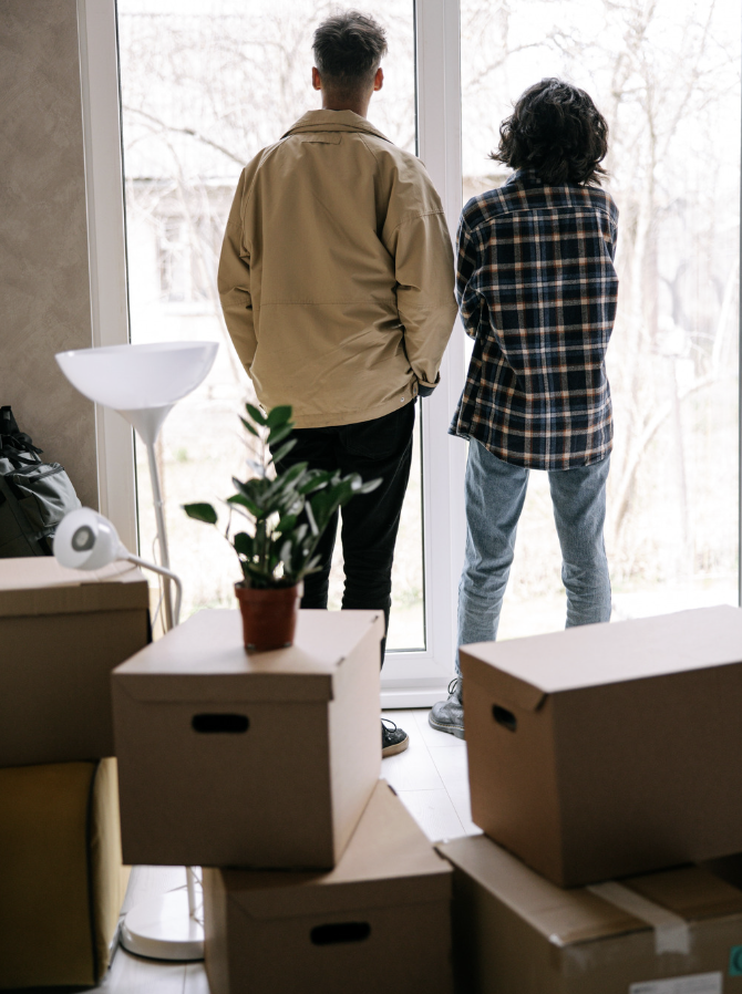 Two people standing near a glass door, looking outside. One is wearing a tan jacket and the other a plaid shirt. The room has moving boxes, a potted plant, and a lamp.