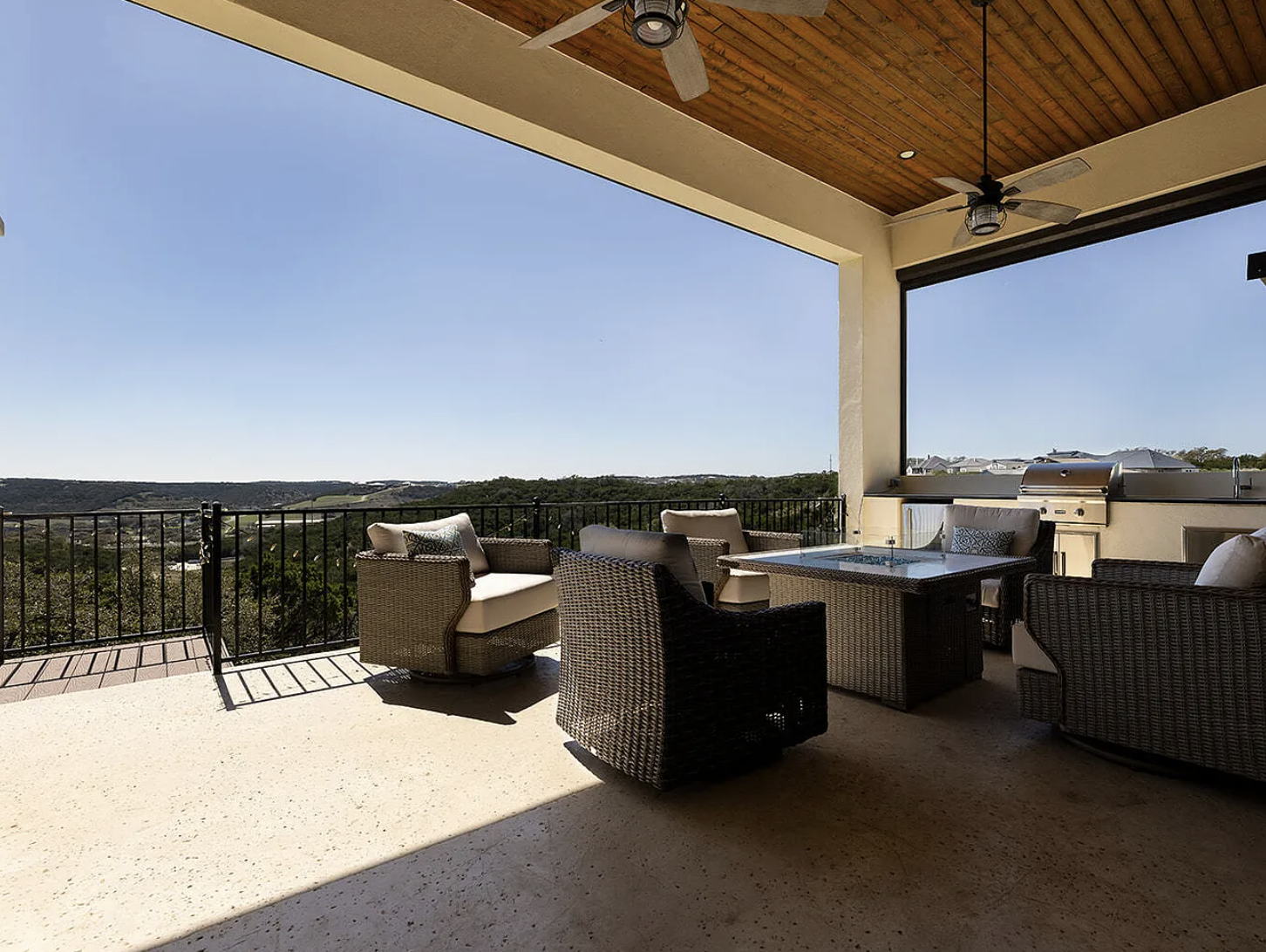 Covered outdoor terrace with wicker furniture, a glass-top table, ceiling fans, and a barbecue grill, overlooking a scenic landscape with trees and distant hills.
