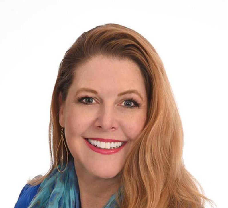 Close-up portrait of a smiling woman with long red hair, wearing a blue top and earrings, against a white background.