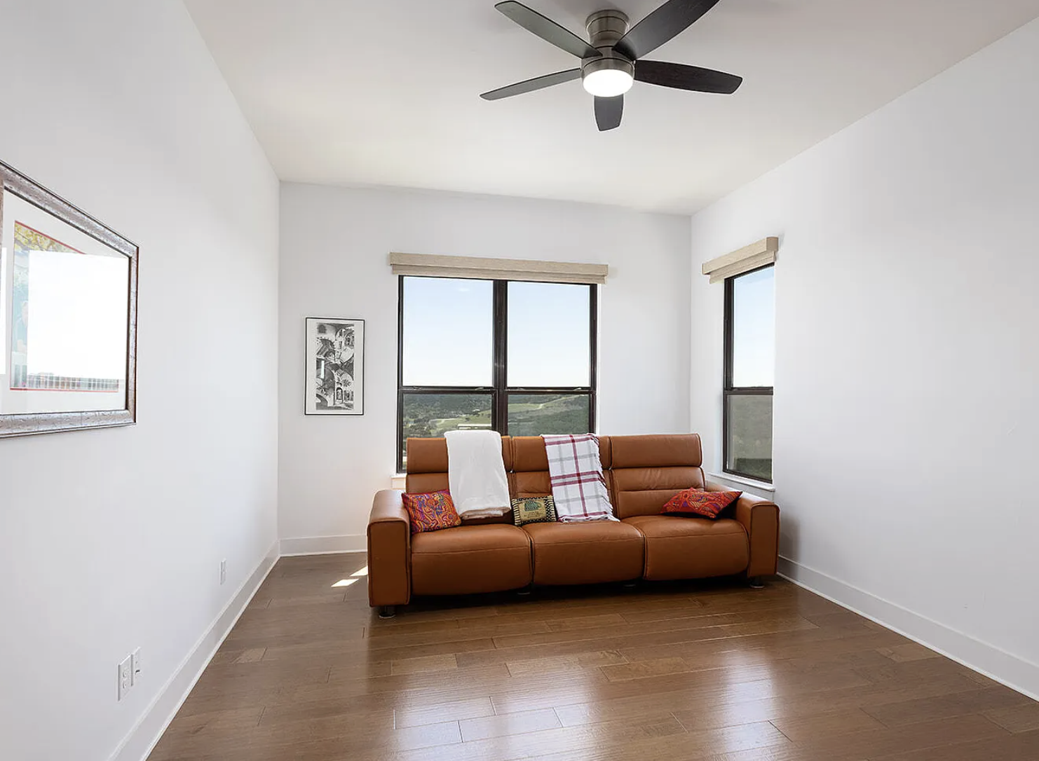 A room with white walls, wooden floor, large windows showing a view of fields and trees, a brown leather sofa with colorful cushions and blankets, framed artwork, a ceiling fan, and beige blinds on the windows.