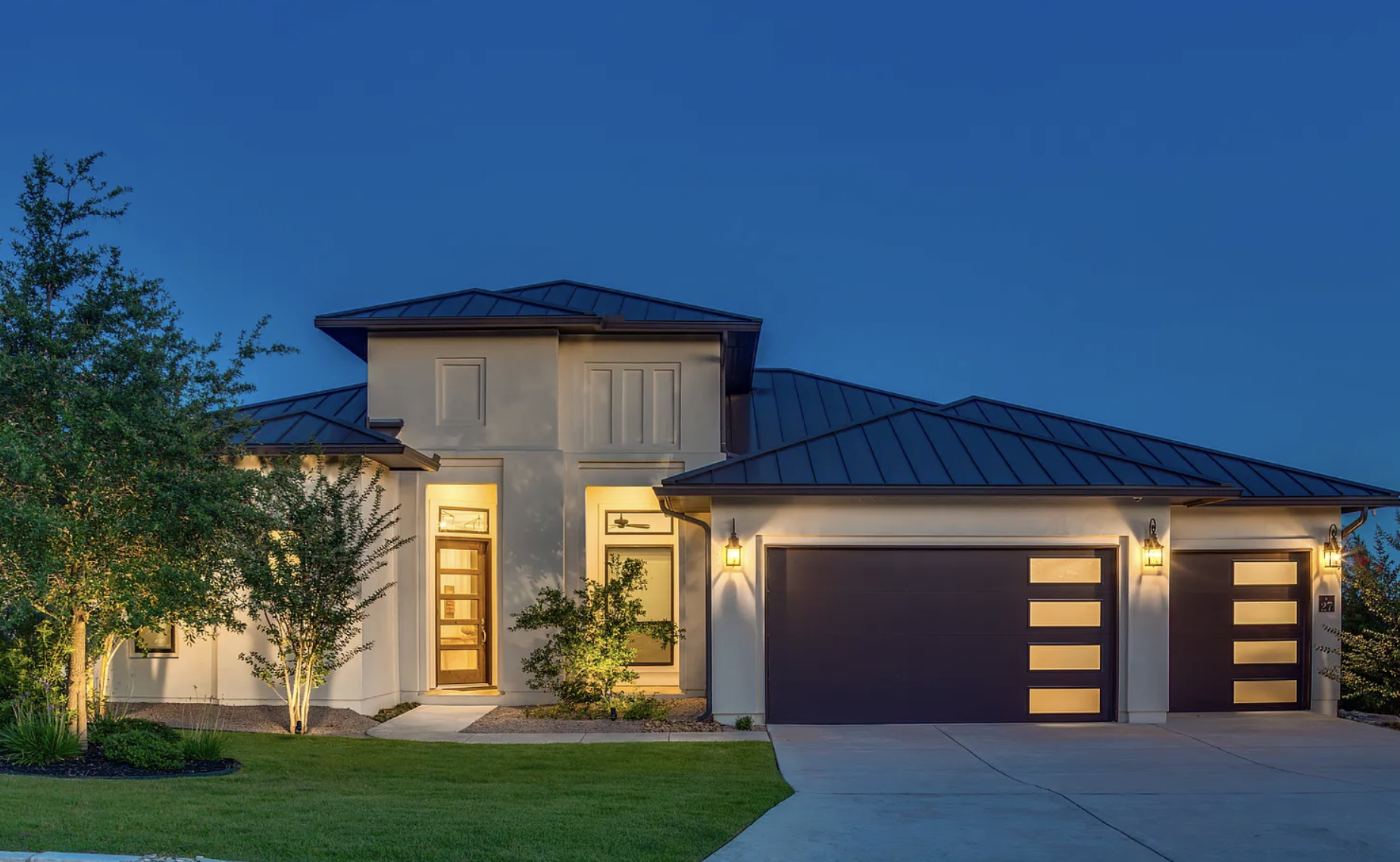 Modern house with a metal roof, illuminated exterior lights, front lawn, trees, and a driveway at dusk of 27 Mariposa Pkwy E., Boerne, Texas