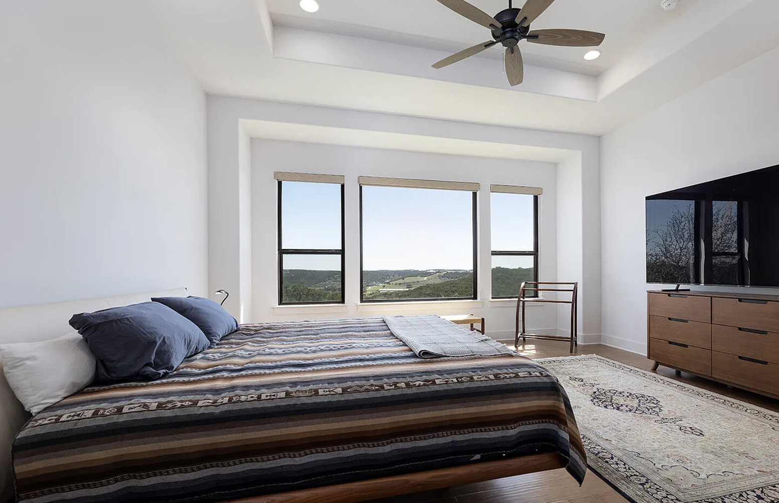 Bright bedroom with large windows overlooking a landscape, bed with striped bedding and navy pillows, wooden dresser with a TV, area rug, ceiling fan, and natural light.