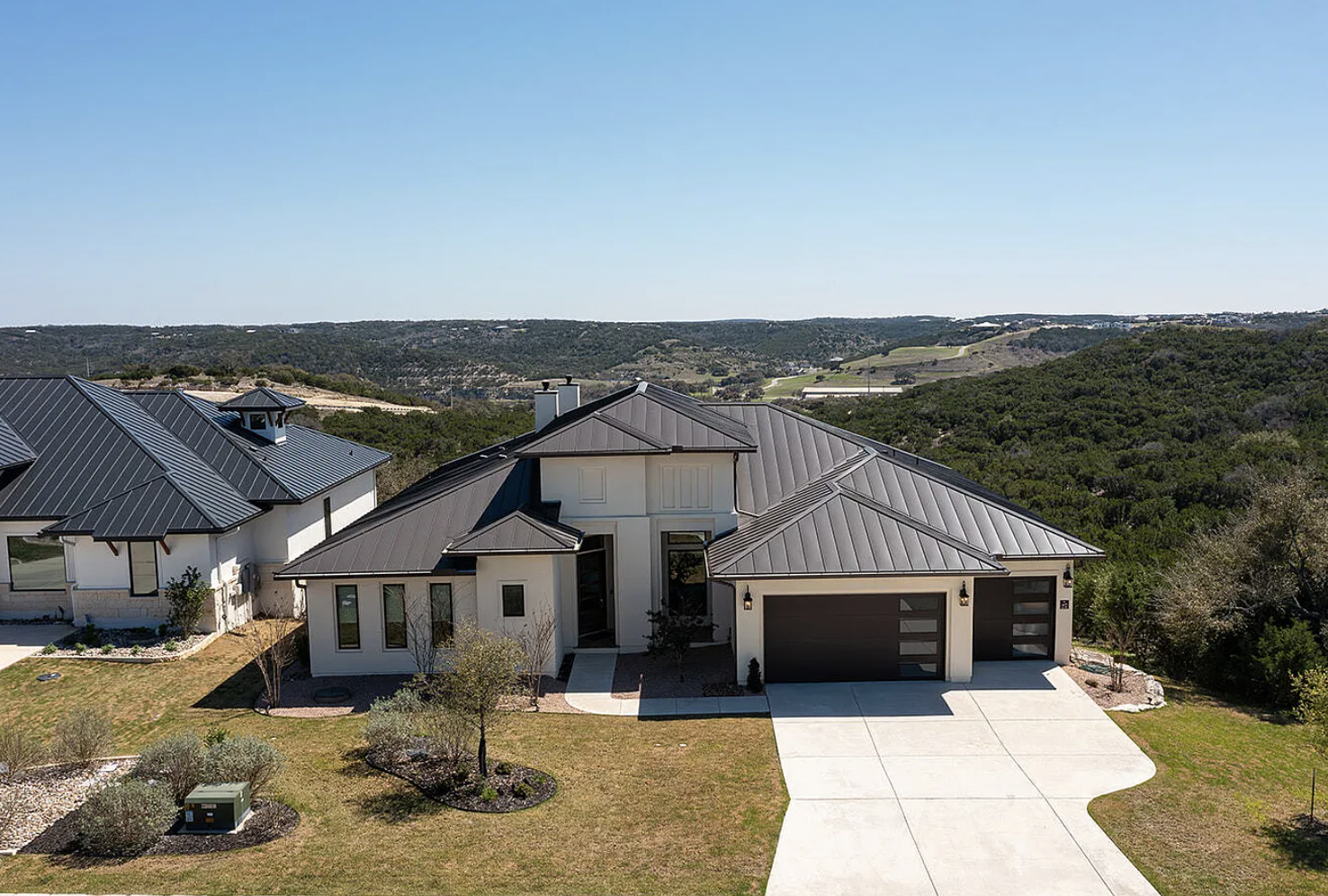 A modern suburban house with a metal roof, white exterior walls, and a two-car garage, surrounded by a well-maintained lawn and small trees, with rolling hills in the background under a clear blue sky.