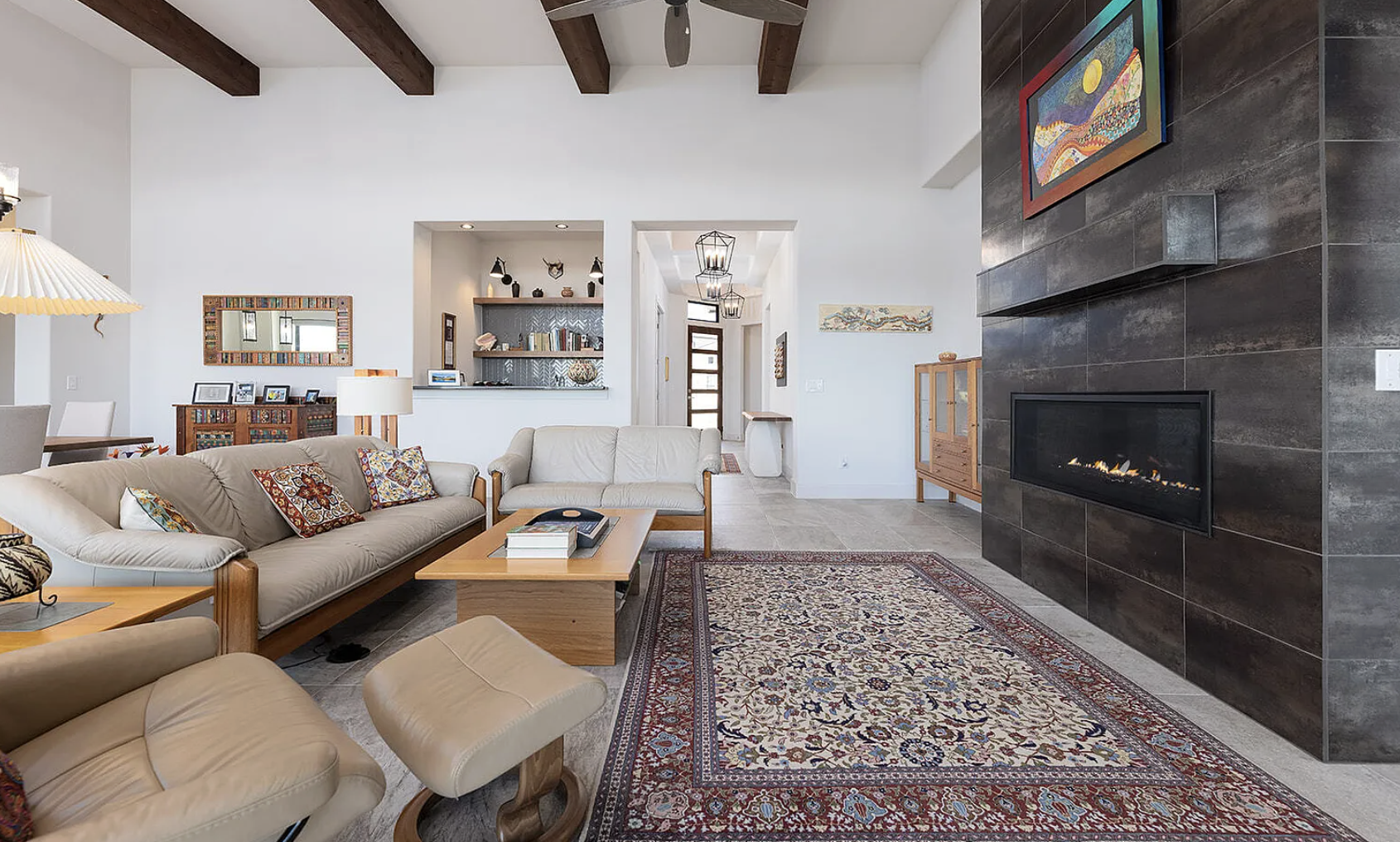 A spacious living room with beige sofas, a patterned area rug, a modern fireplace with dark tile surround, and wood beam ceiling. The room has white walls, decorative art, and built-in shelves with books.
