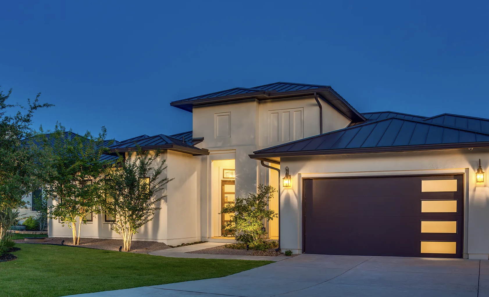 Modern house with white exterior, dark garage door with window panels, landscaped front yard with trees and grass, illuminated by front porch and wall lights at dusk of 27 Mariposa Pkwy E., Boerne, Texas