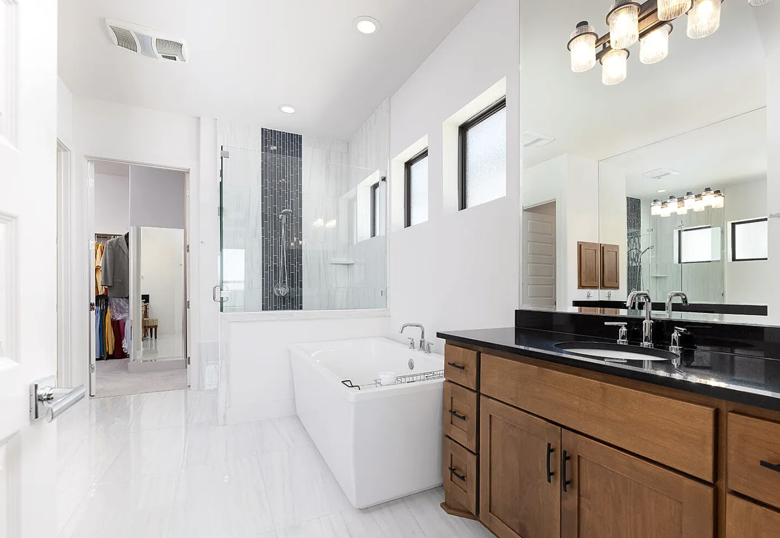 Bathroom with black countertop vanity, wooden cabinets, a large mirror, a bathtub, a glass-enclosed shower with gray and black tiles, four small windows, and a walk-in closet visible through an open door.