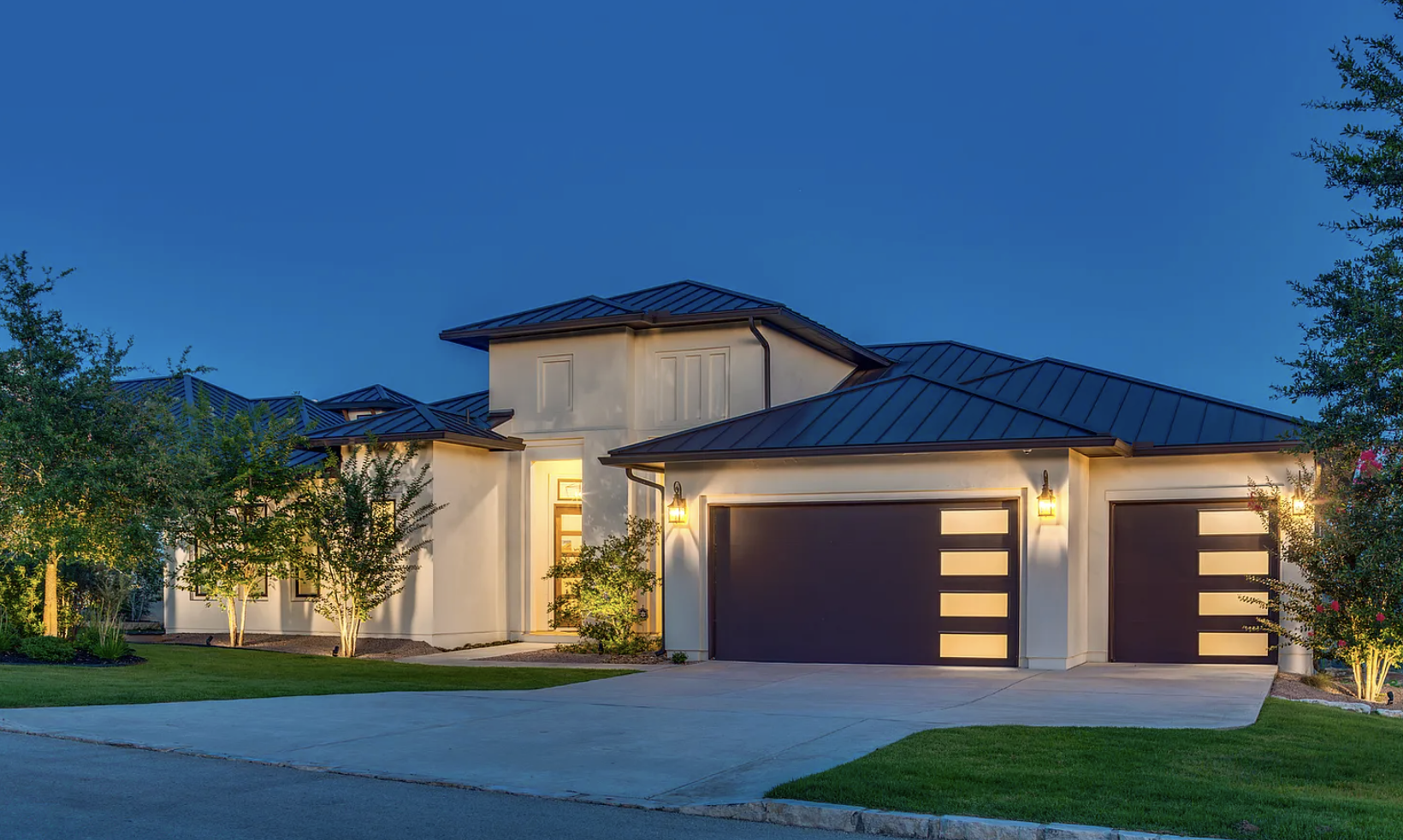 A modern two-story house with a dark metal roof, illuminated exterior lights, dark garage doors with rectangular windows, a front door with glass panels, well-lit landscaping, and a manicured lawn at dusk.