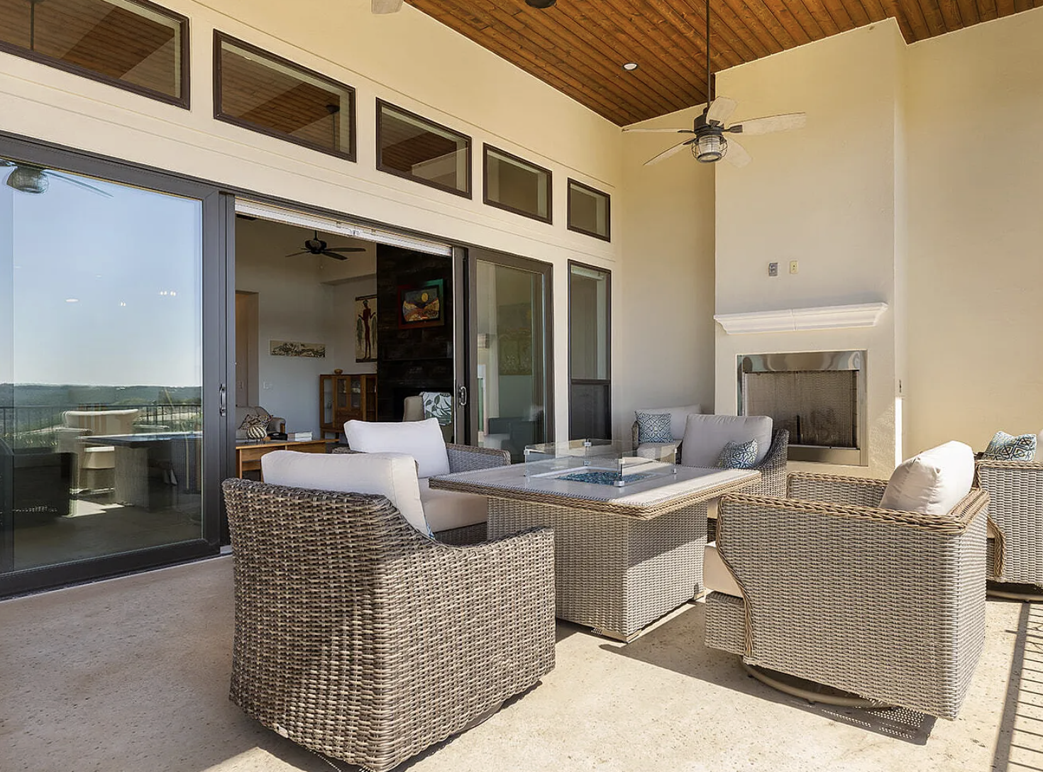 Outdoor patio area with wicker chairs, cushions, a table with a glass display, and a fireplace, overlooking a clear sky and cityscape through glass sliding doors.