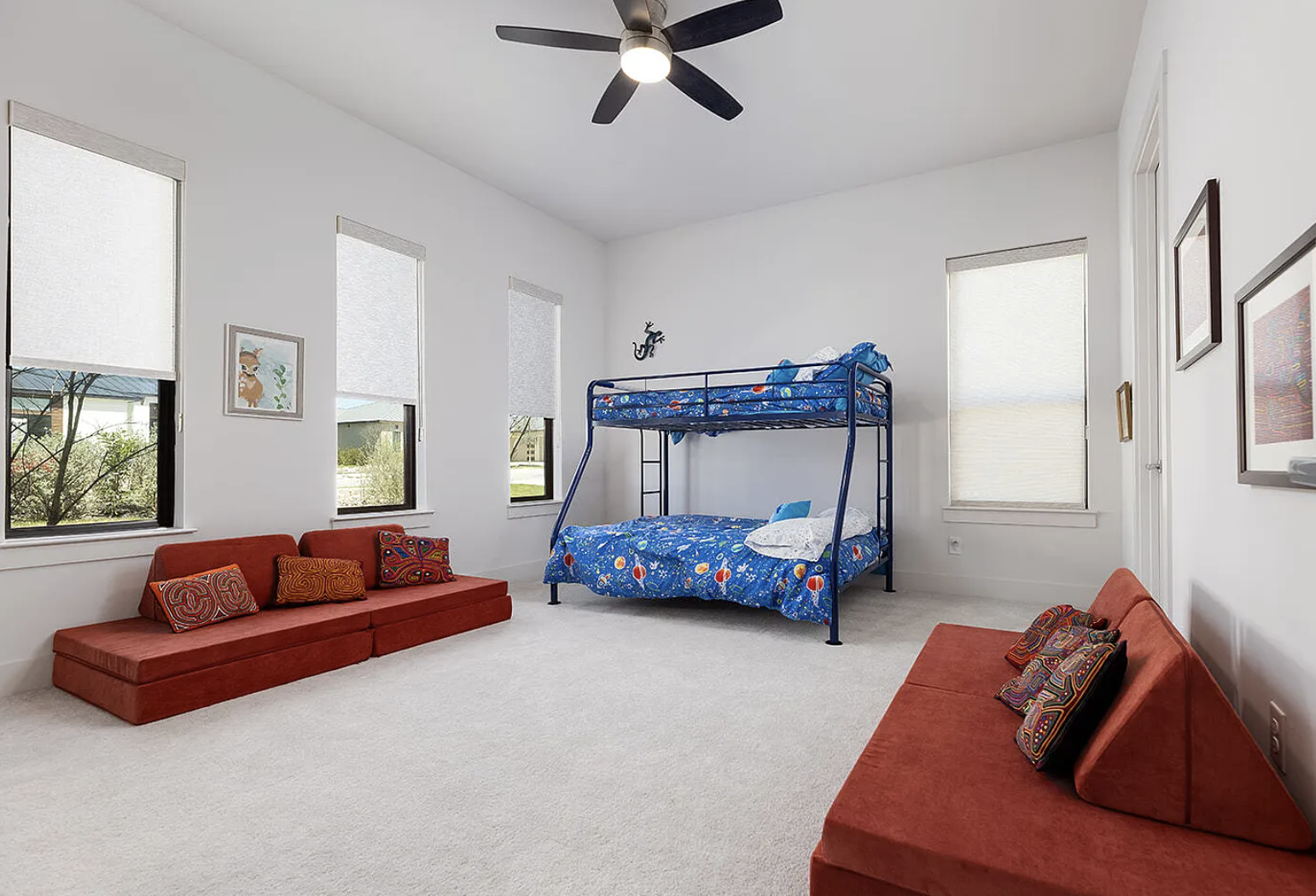 A bright children’s bedroom with white walls, three windows with white blinds, a ceiling fan, a blue bunk bed with space-themed bedding, two red sofas with colorful pillows, and framed artwork on the walls.