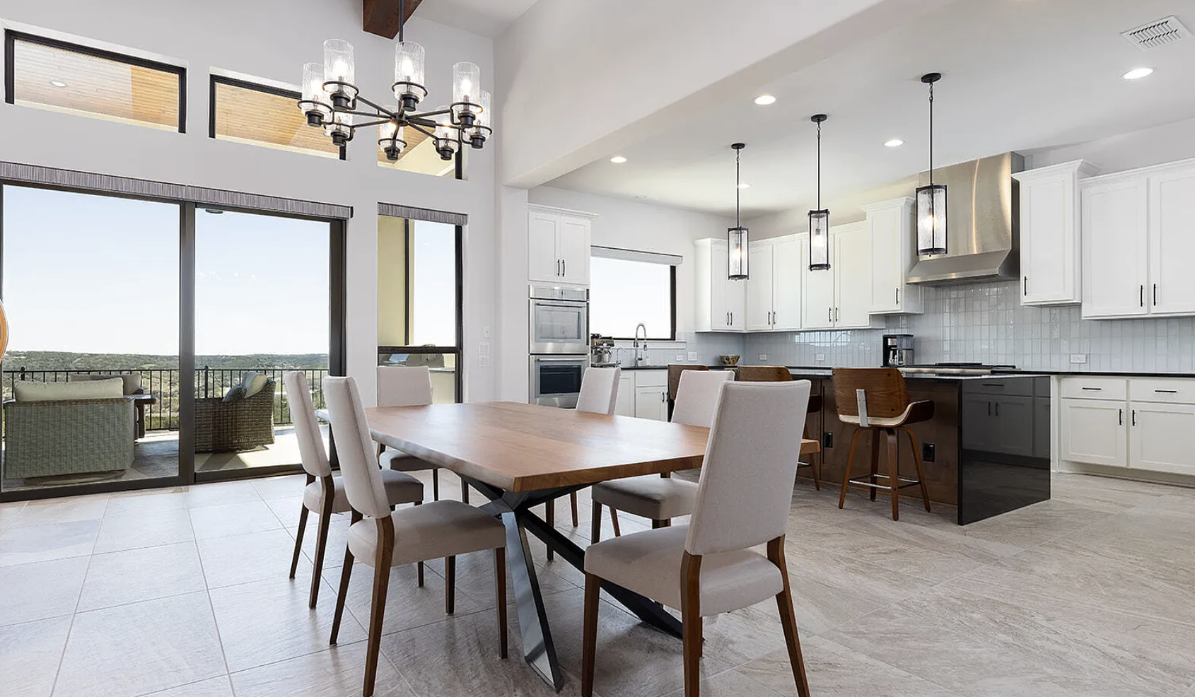 Modern kitchen and dining area with a wooden table, white chairs, and large sliding glass doors leading to an outdoor seating area with a view. White cabinets, stainless steel appliances, and pendant lighting are visible.