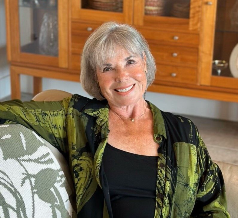An elderly woman with short gray hair smiling and sitting on a sofa in a room with wooden cabinets in the background.