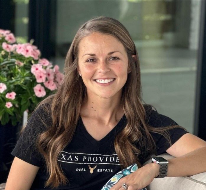 A young woman with long wavy brown hair, wearing a black t-shirt with 'TEXAS PROVIDE REAL ESTATE' written on it, smiling at the camera, seated outdoors with a pot of pink flowers and a glass door in the background.