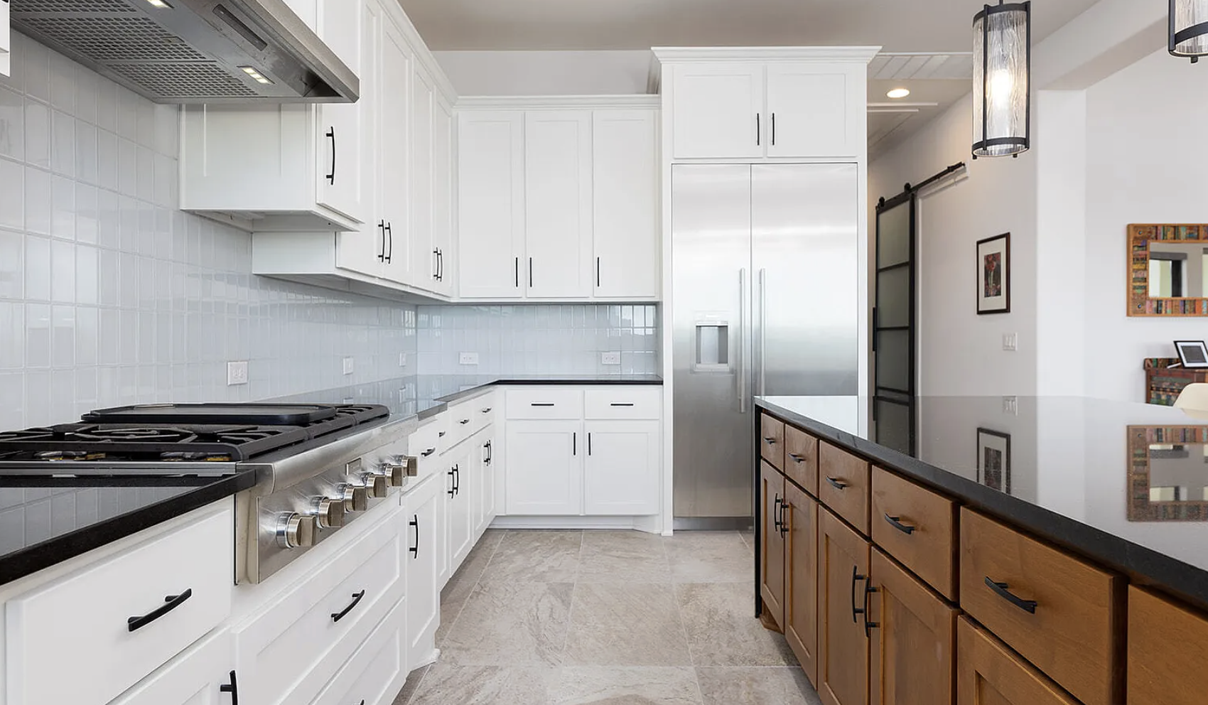 Modern kitchen with white upper and lower cabinets, a black countertop, a large stainless steel refrigerator, a stainless steel stove, a black countertop, and a wooden island with drawers. There is a tiled backsplash and neutral tile flooring, with d