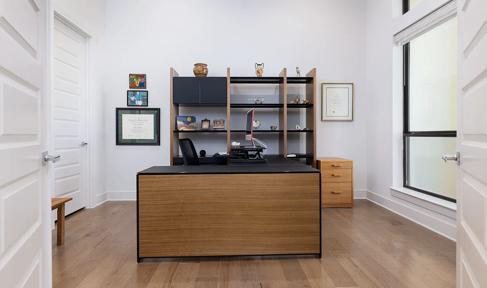 Empty office with a wooden desk, black countertop, computer, black chair, wooden shelves holding decorative items and framed certificates, wooden drawers, large window with frosted glass, hardwood floor, white walls, viewed from doorway.