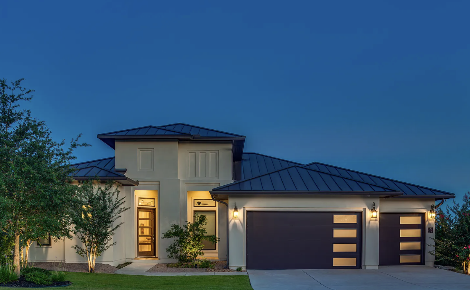 A modern two-story house with a dark-colored roof, well-lit exterior, a double garage with dark doors, and a small front yard with trees and grass, during twilight.
