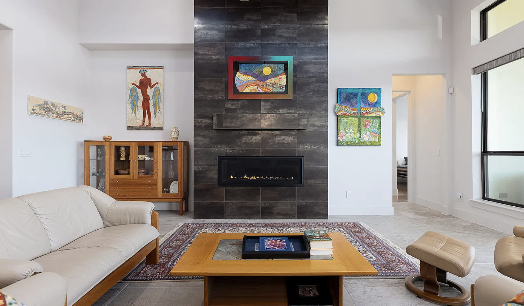 Living room with a beige sofa, wooden coffee table with books and artwork, colorful paintings on the white walls, a modern fireplace on a dark tiled wall, a wooden display cabinet, and large windows letting in natural light.