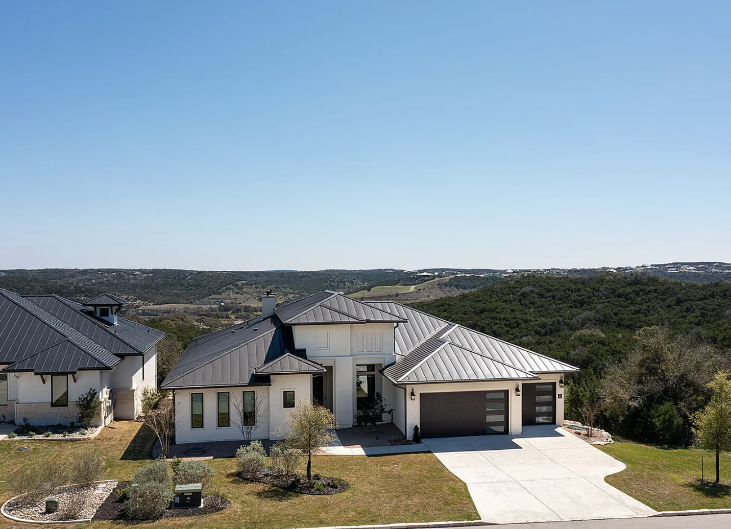 Modern house with white exterior, gray metal roof, and two-car garage, located in a suburban neighborhood with landscaped yard and surrounding hills in the background under clear blue sky.