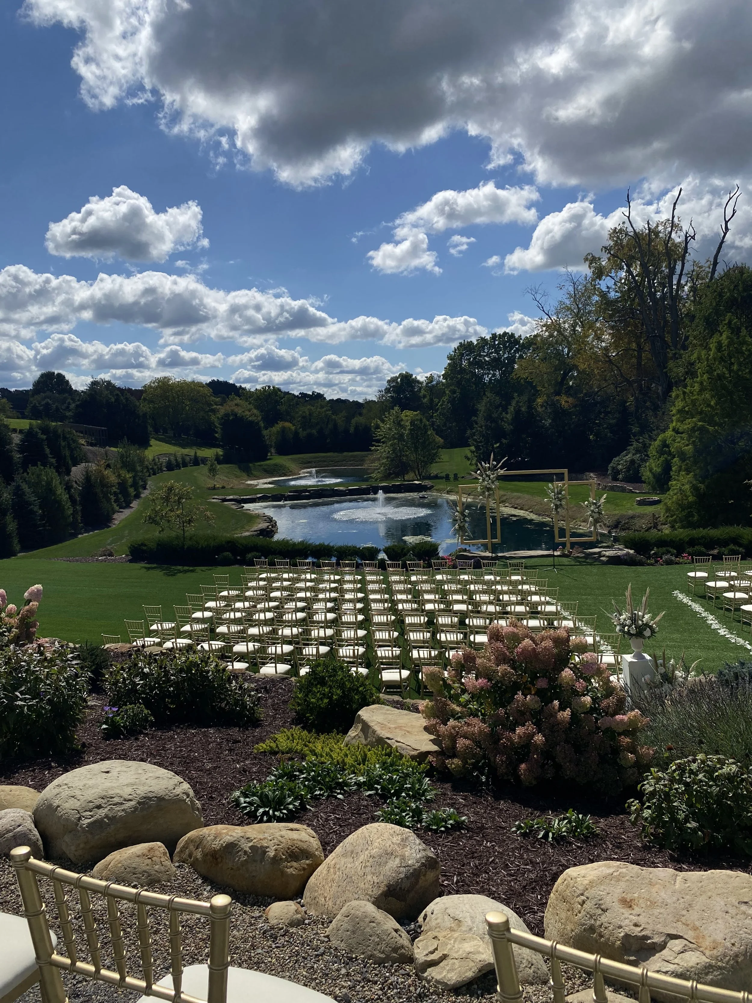 An outdoor wedding setup on a grassy field near a pond with chairs arranged facing an altar, surrounded by flowers and scenery including trees, rocks, and a cloudy sky.