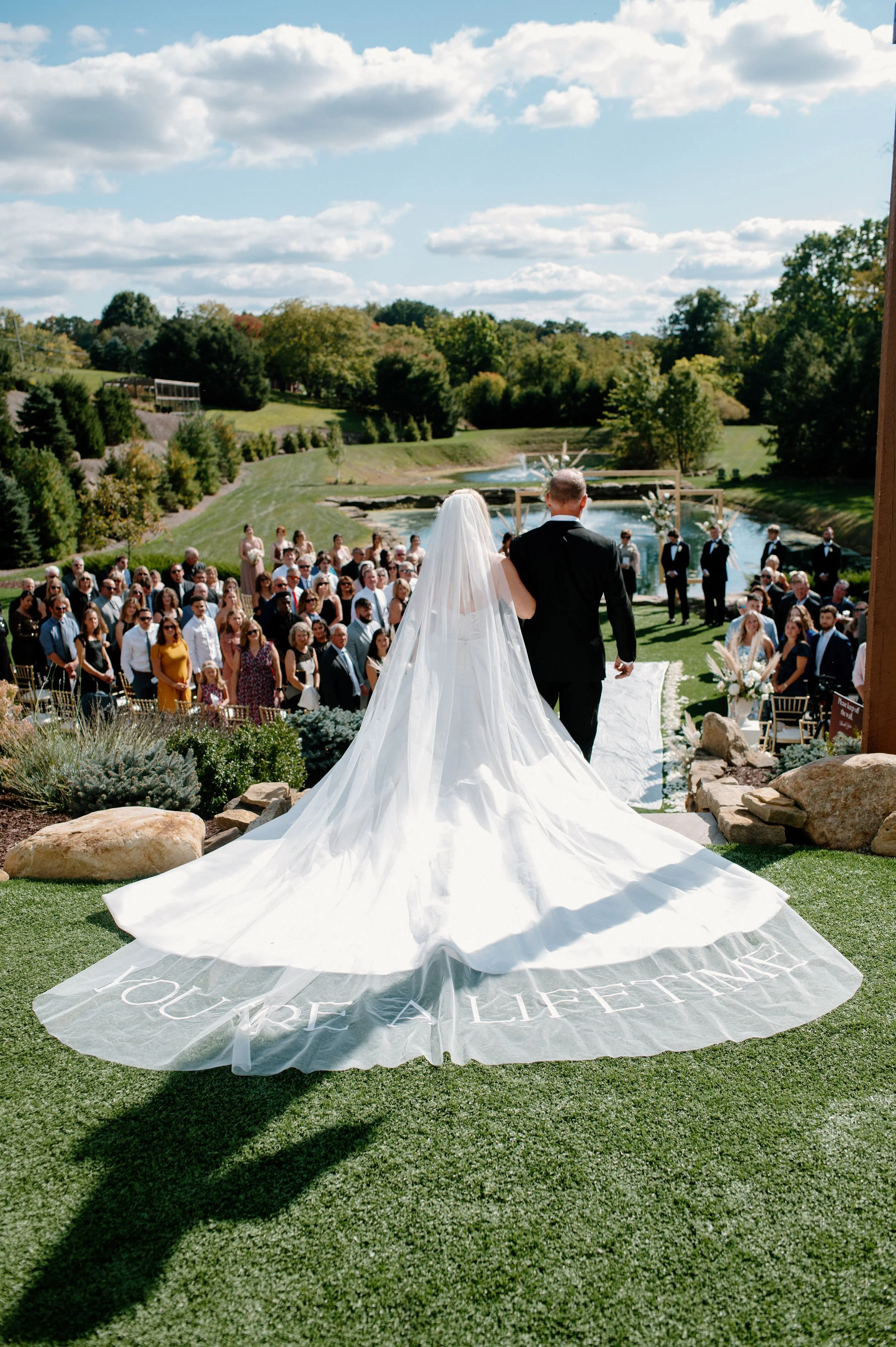 Bride and groom walking down a wedding aisle toward guests in an outdoor landscaped setting with a pond, trees, and blue sky.