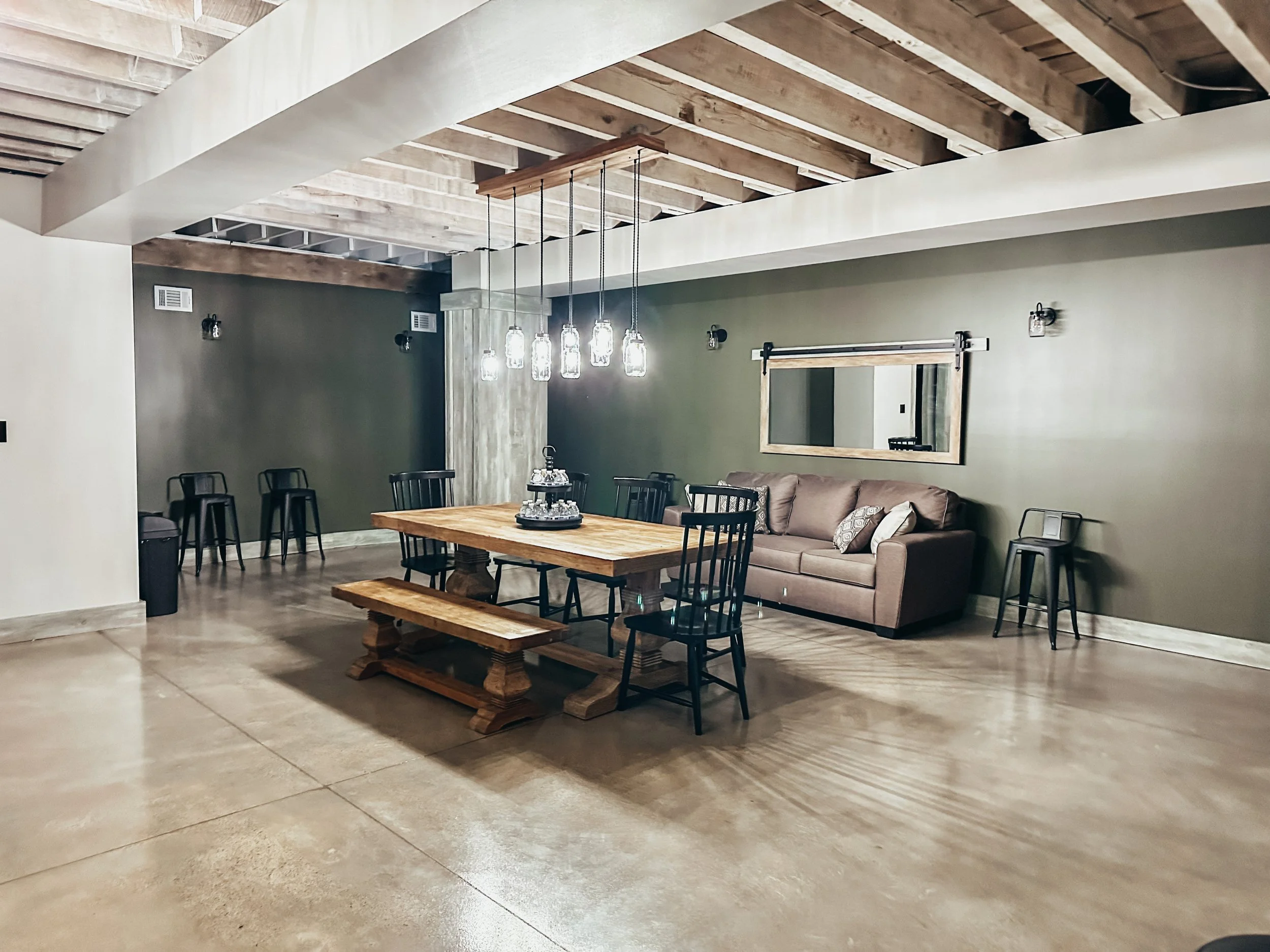 Spacious ready room with polished concrete floor, wooden ceiling beams, and light green and white walls. Contains a large wooden dining table with black chairs, a beige sofa, a large mirror, pendant lights, and several black barstools.